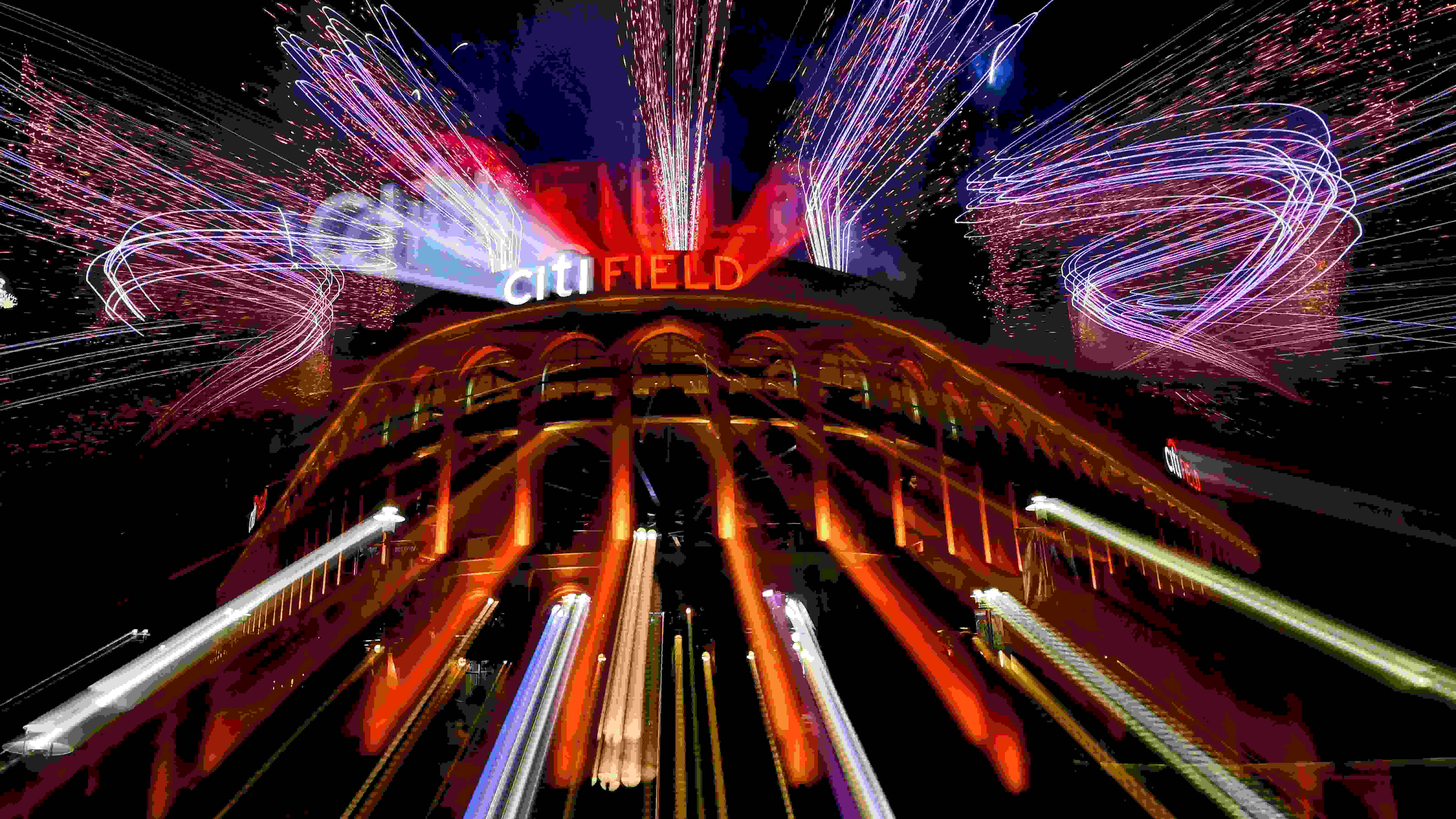 A photograph of Citi Field, the home stadium of the New York Mets, at night. There are bright, colorful lights and fireworks going off.