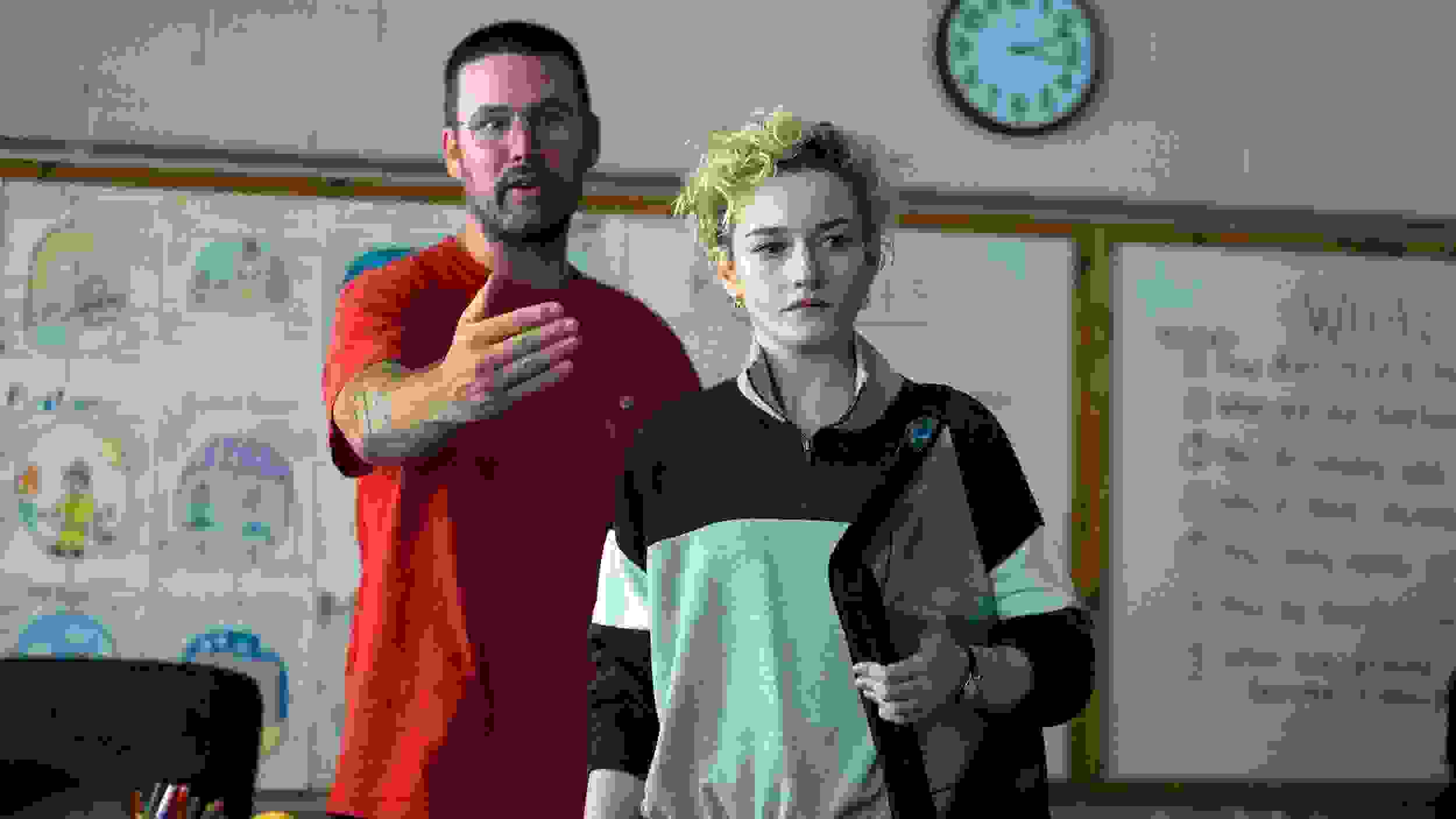 A person in a red shirt gestures in a classroom, appearing explanatory, while another with short curly hair listens intently. A whiteboard and clock are in the background.