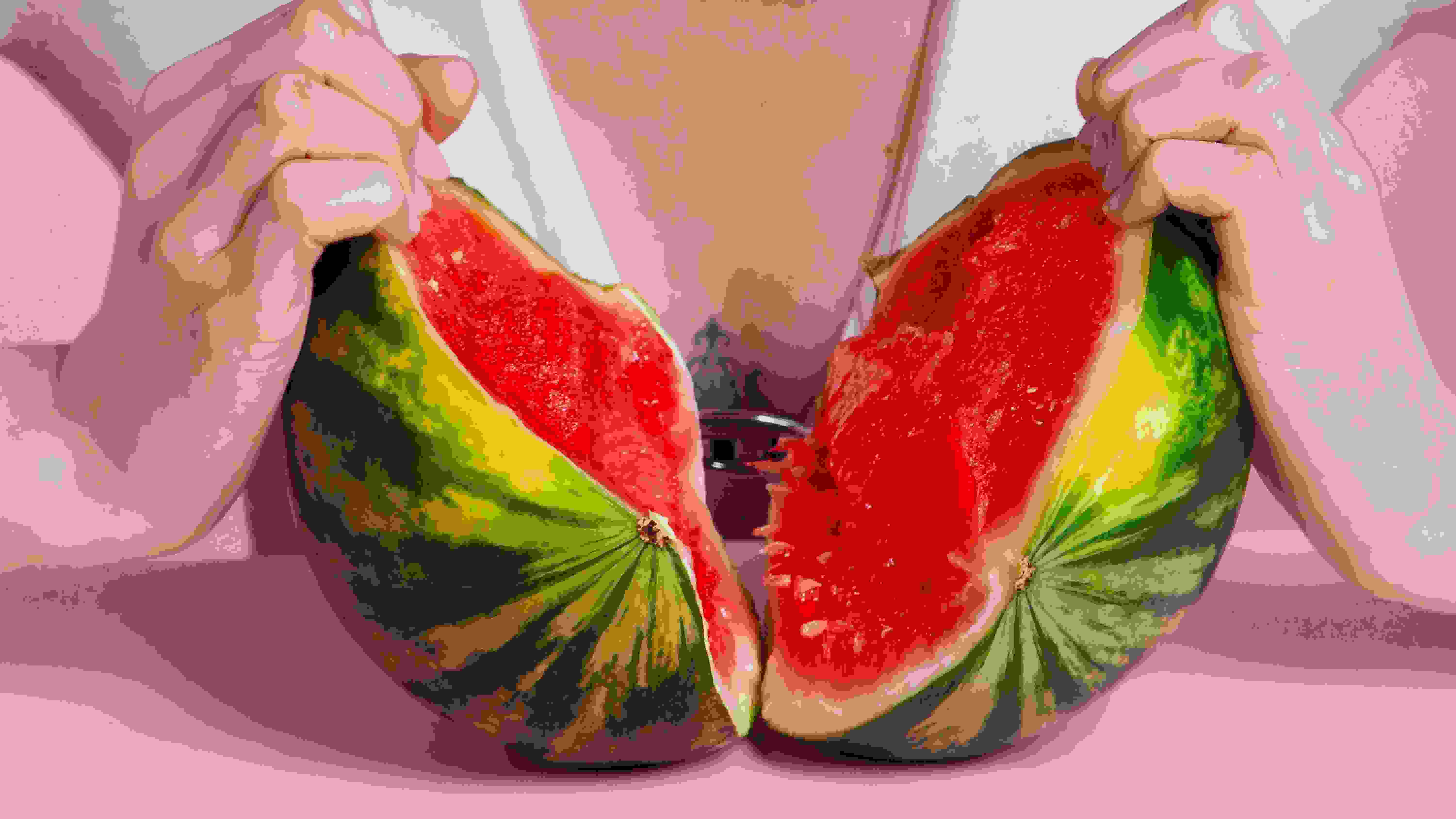 Two hards ripping open a small watermelon in front of a person's chest. There's a pink wall and table in the image as well.
