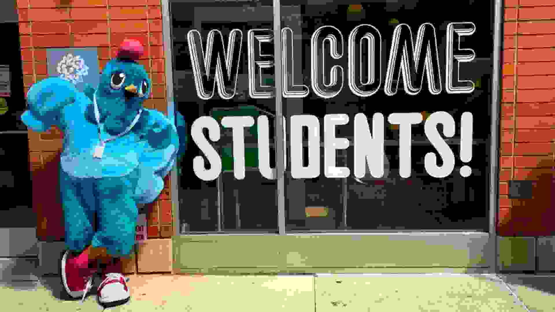 A bright blue pigeon mascot leans against a brick wall next to a window with "Welcome Students" on it in big letters.