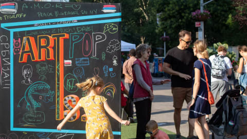 An image of the black chalkboard box with the words "Art Is..." with writings and drawings in chalk from the public. A family enjoying Salsa and Art Nights on the Flatiron North Plaza. 