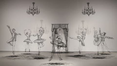 An installation photographs of a set of interrelated sculptures which together depict a ballet performance—six dancers, two chandeliers and a set of curtains. The sculptures are done in wire so they look like line drawings.