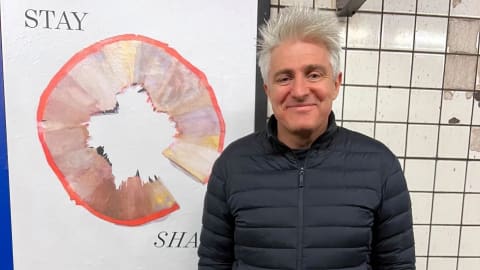 A man with gray hair, wearing a black jacket, smiling as he stands next to a poster in the subway. 