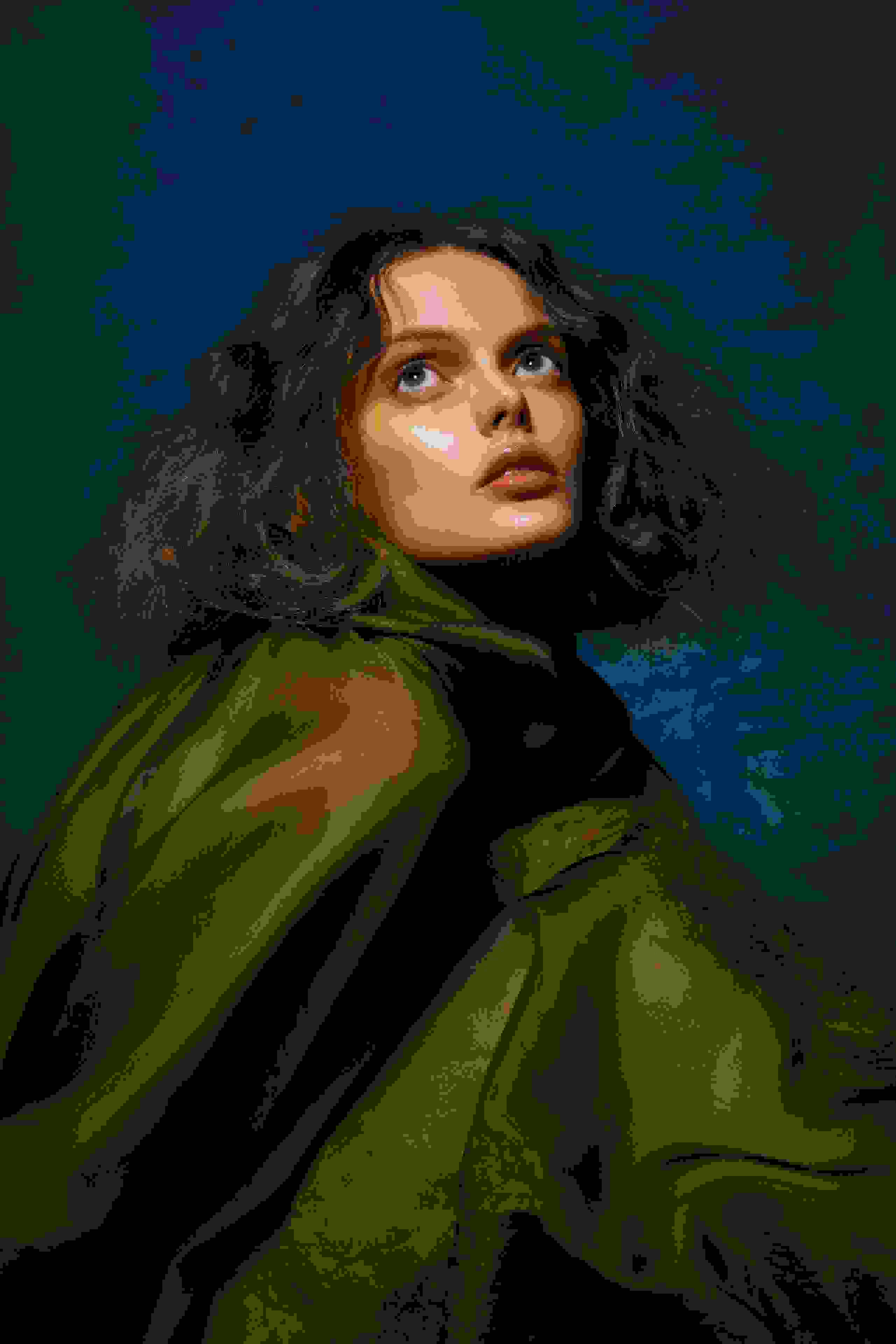 A fashion portrait of a woman against a blue background. She has shoulder length, wavy hair, is wearing a sheer green drapery, and looking up and off camera.