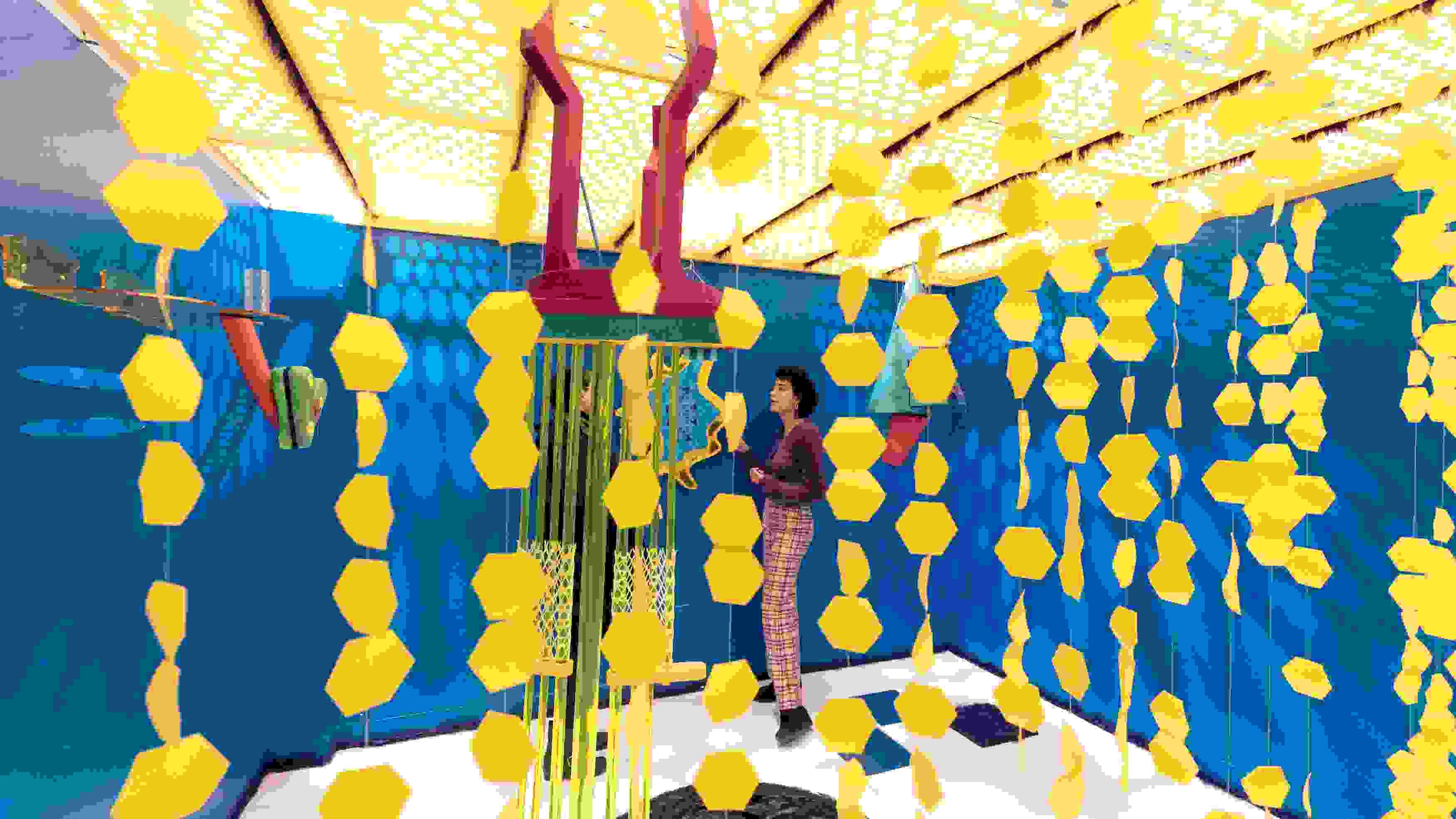 Person standing in an art installation with bright blue walls and mobiles of yellow hexagon-like shapes.