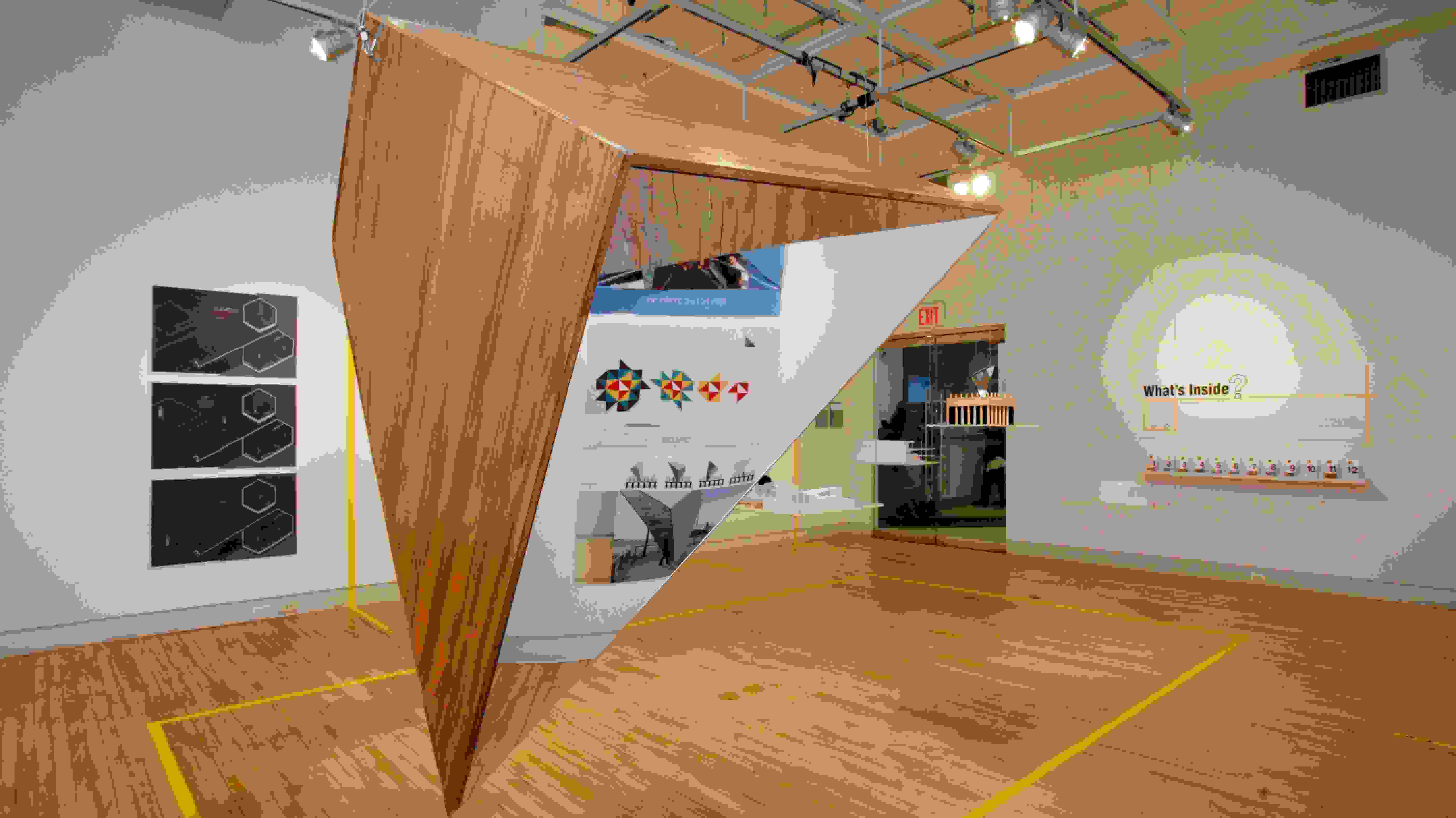 Mirrored pyramid sculpture hangs from the ceiling in the center of the gallery. There are prints on the wall behind the sculpture. On the wall to the right of the sculpture reads, "What's Inside?"