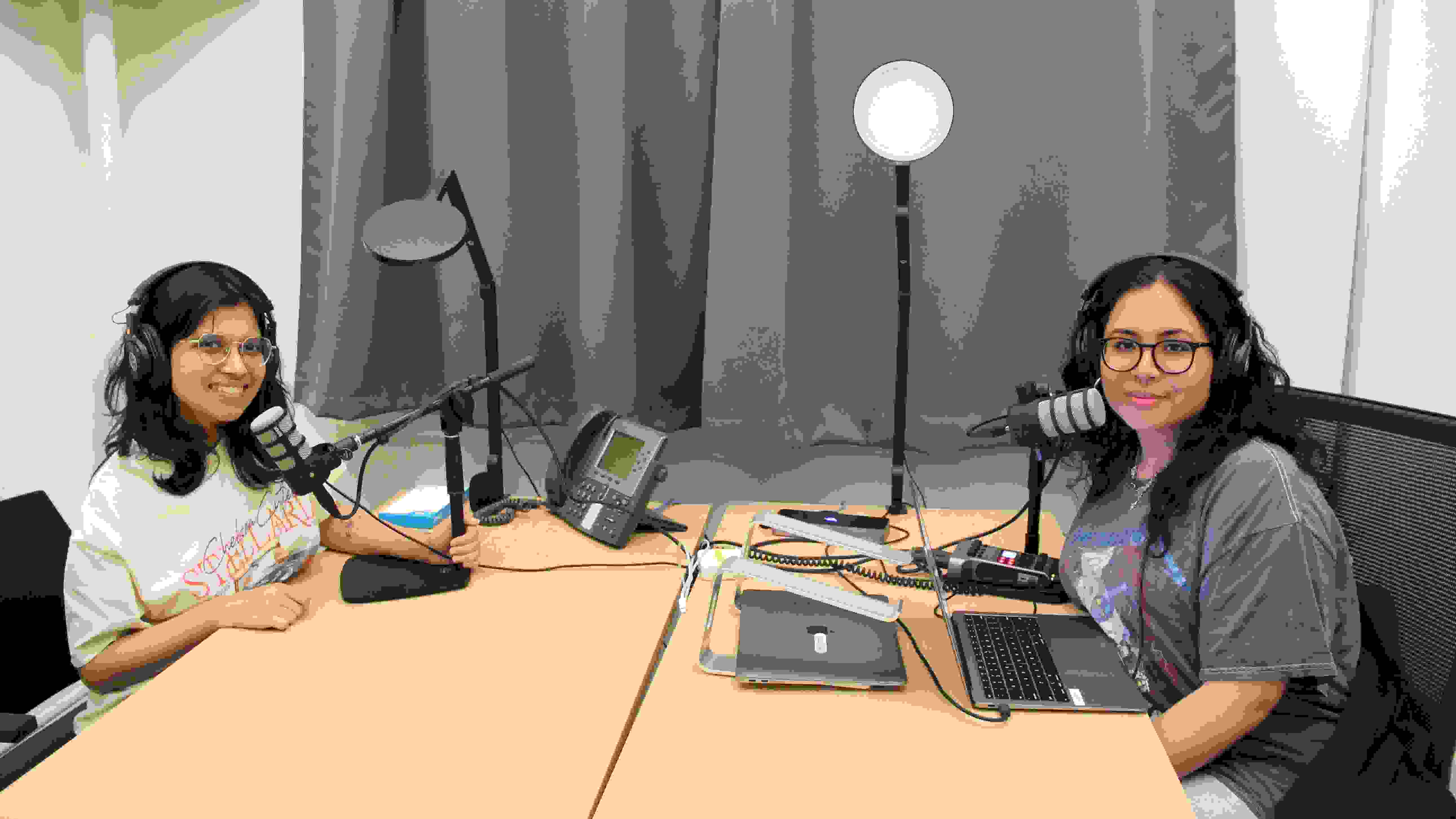 Two people sit at a long opposite one another smiling for the camera; they are using podcast production equipment such as microphones, laptops, headphones and lights.