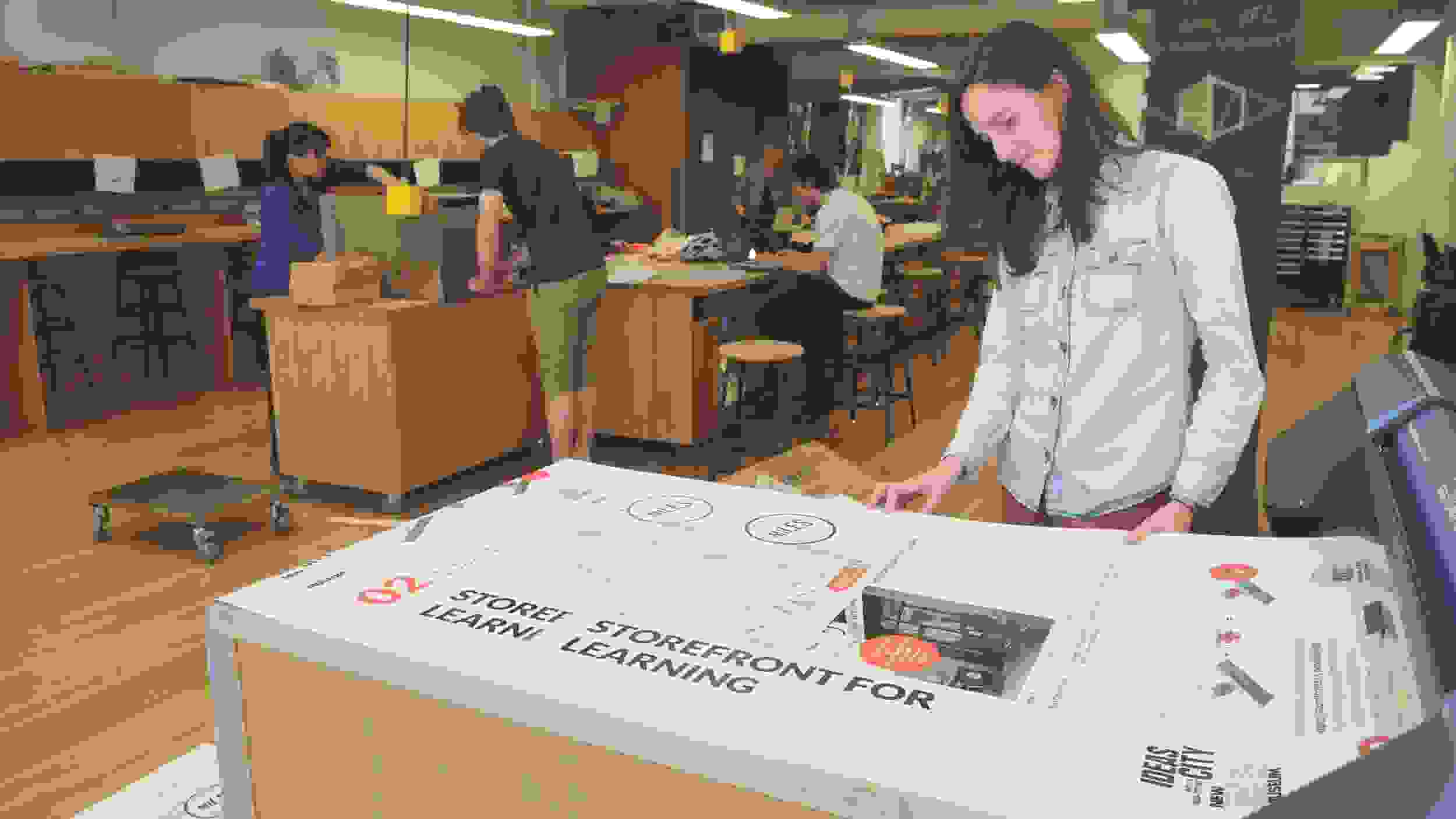 Wide view of a maker space with wooden floors, cabinetry, and workspaces. In the foreground, a student looks on as their project is being printed from a large printer.