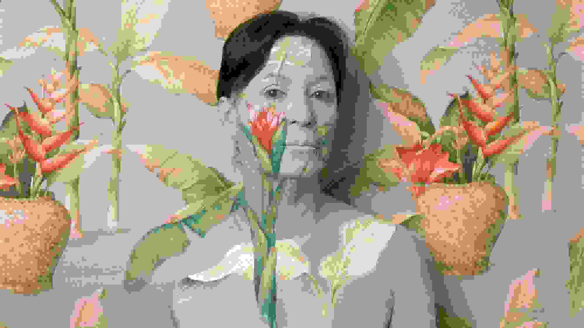 A photograph of a woman from the chest up standing in front of an ornately-wallpapered wall. The wallpaper is pale blue, and there are green plants with orange flowers sticking out of woven pots. The woman is painted with the same pattern to blend into the wallpaper, with only her brown hair standing out.