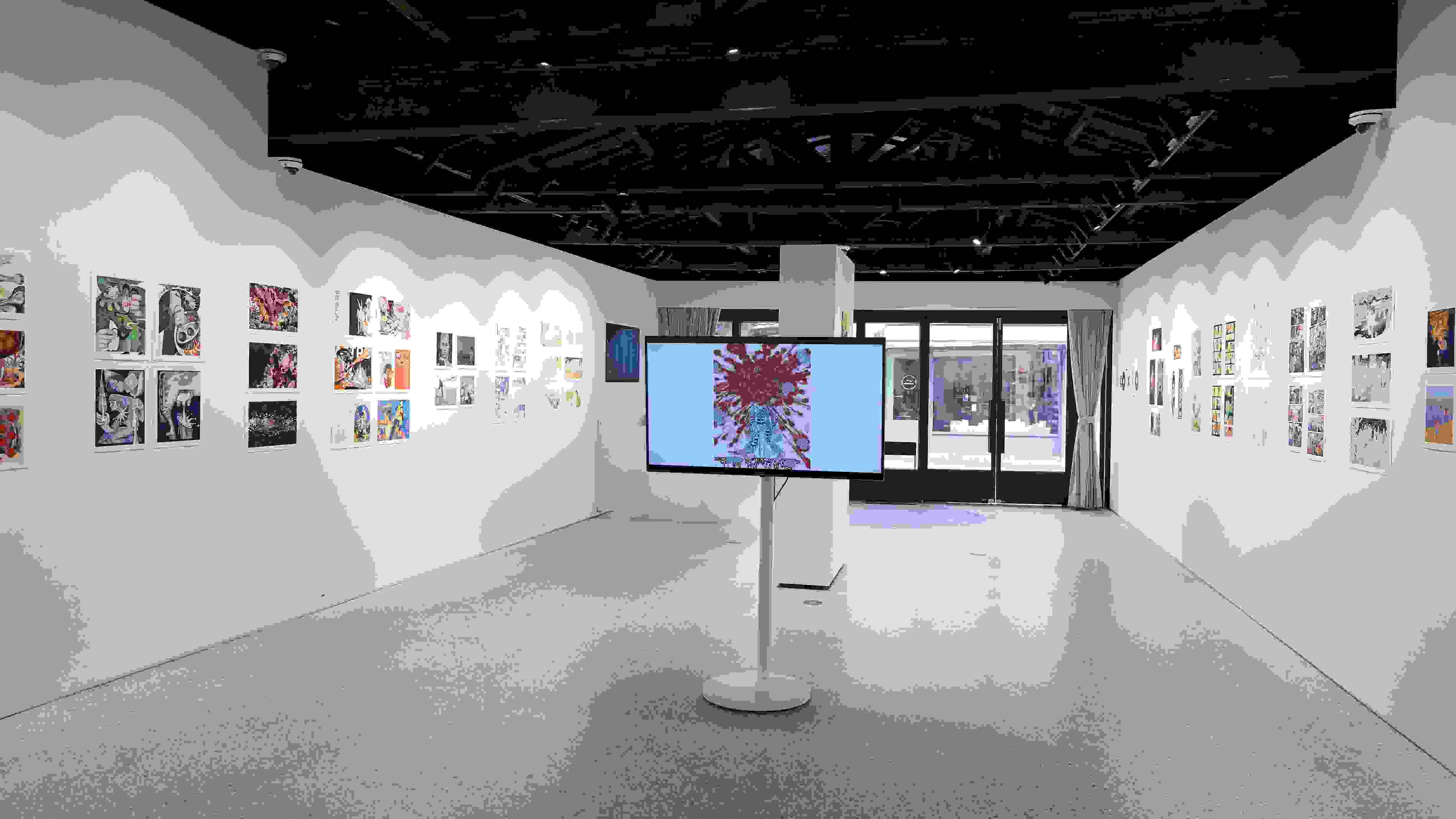 Gallery installation displaying colorful comic and illustration-style artworks arranged linearly along both walls. A large monitor on a white pedestal at the center shows a figure with red abstract patterns. The space features an open ceiling with exposed dark wooden beams, track lighting, and natural light filtering through glass doors at the far end.