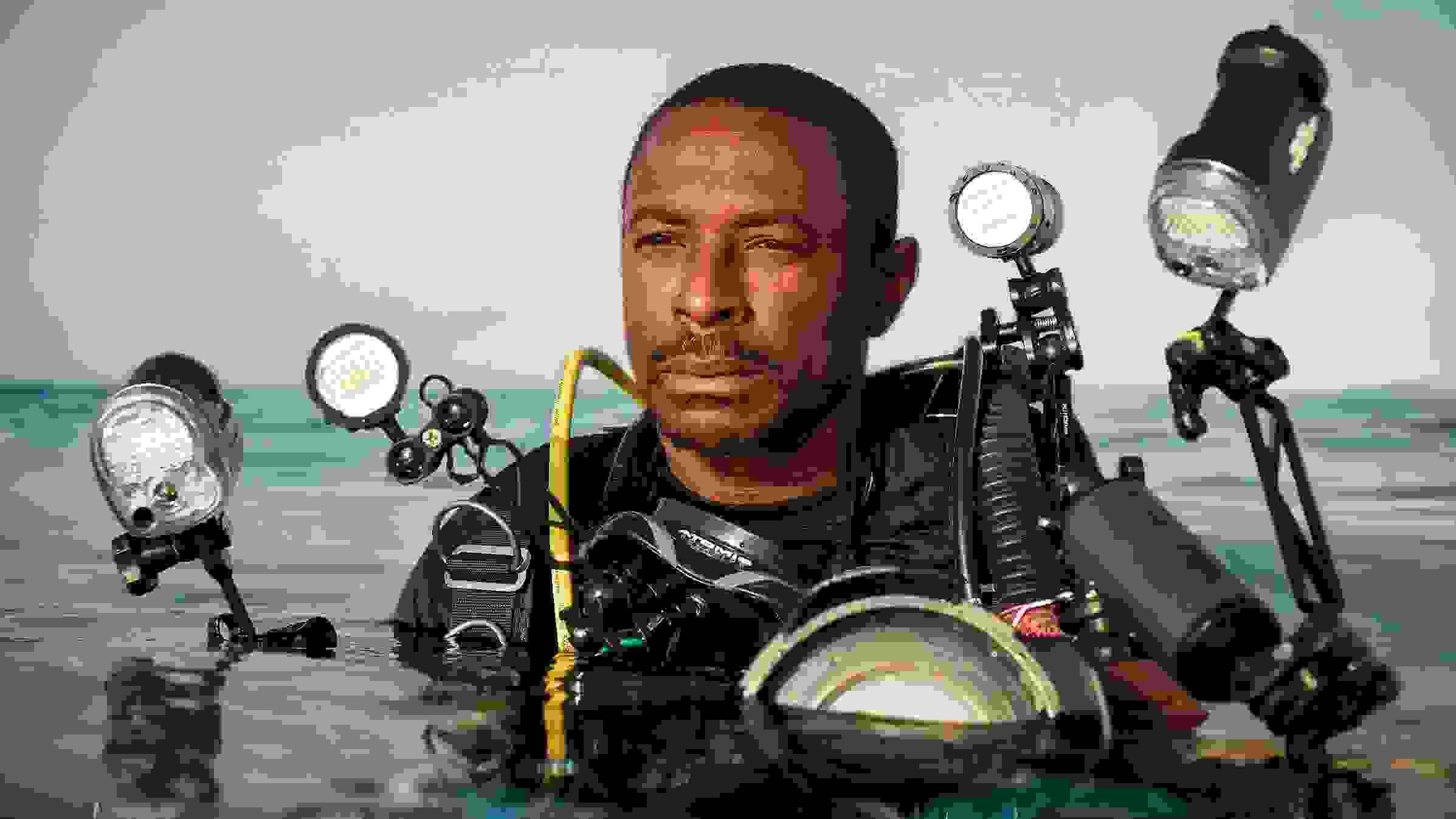 A portrait of a scuba diver in water. He has darker skin and short hair, is wearing a black top, and is equipped with numerous lighting and camera equipments strapped to his body. His body is submerged in water from the chest down.
