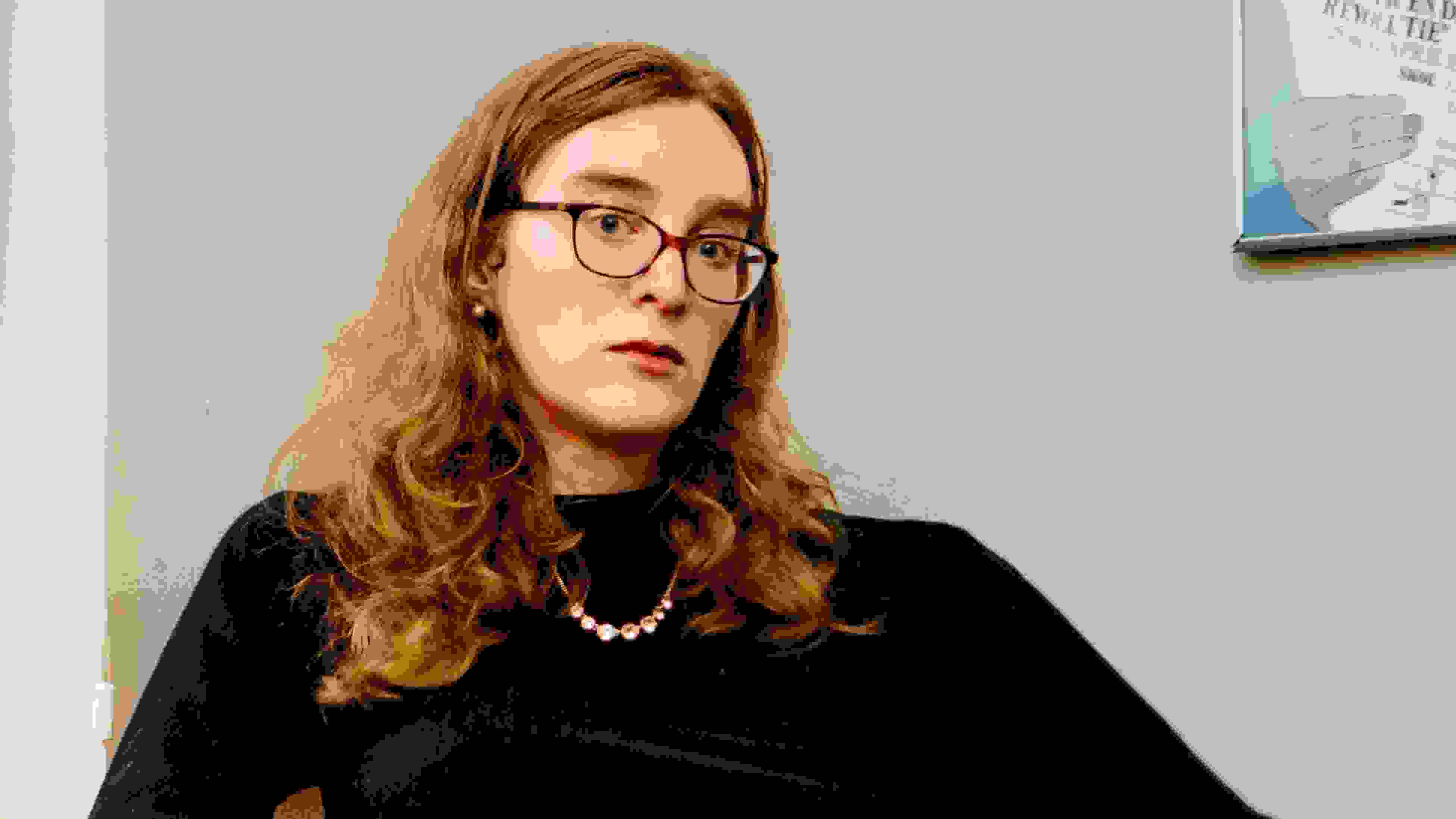 Photo portrait of the writer Grace Byron, sitting in a chair in a black turtleneck with her hands placed on top of one another. Grace wears glasses and looks directly at the lens.