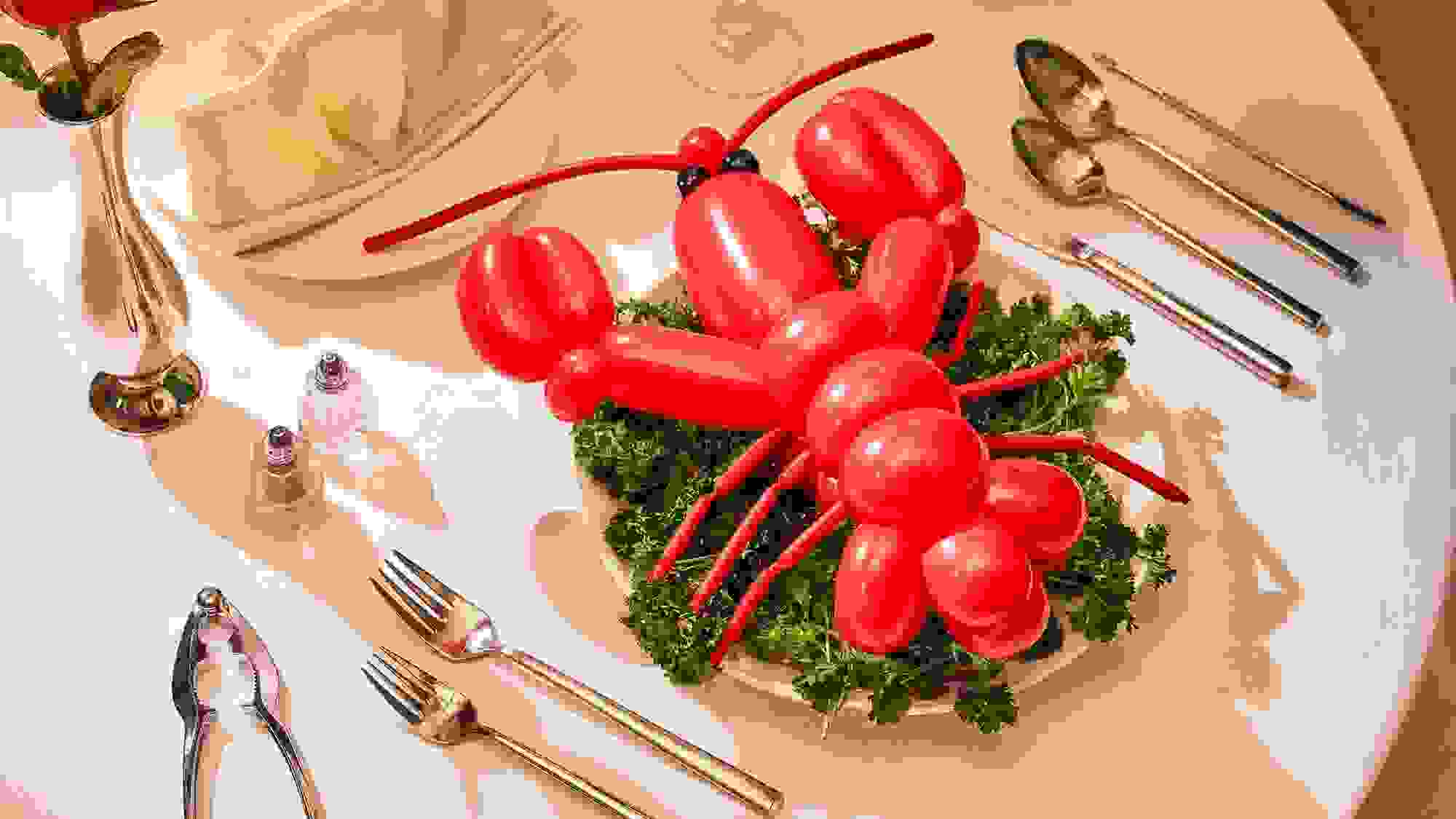 A photograph of a lobster shaped red balloon sitting on top of a bed of kale leaves on a dinner plate. Surrounding the scene is a dinner table fully set up with cutlery,  a dinner napkin folded into a fan, and a single rose in a tin vase.