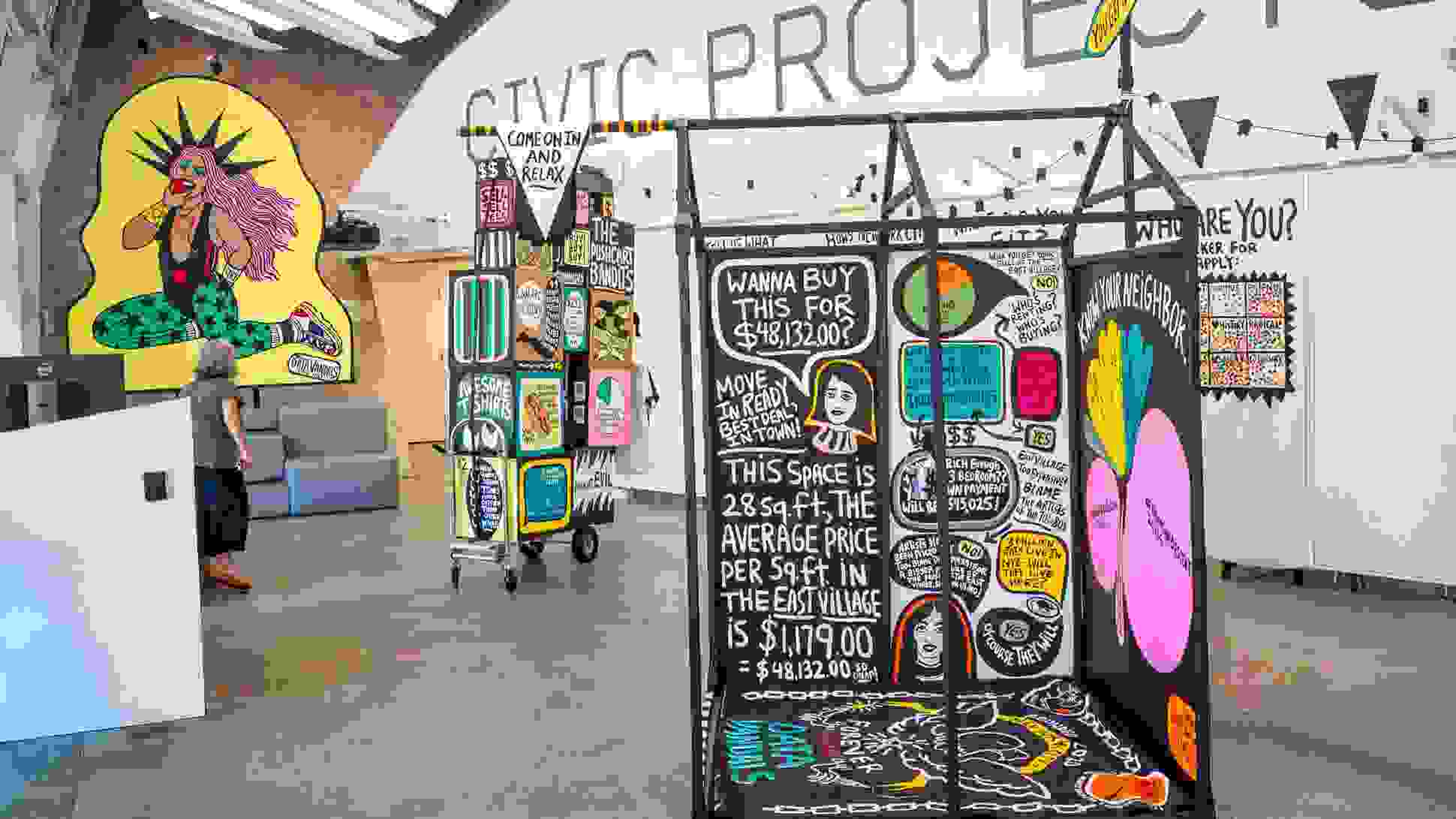 Interactive data visualization installation in a modern gallery space featuring colorful, street art-inspired panels with housing cost data. The central black panel displays NYC housing statistics including '$48,132.00' and text about East Village rental prices per square foot. Surrounding panels feature vibrant graphics with pink, yellow, and green elements. A person in casual clothing examines the display while standing on polished concrete floors under industrial ceiling architecture.