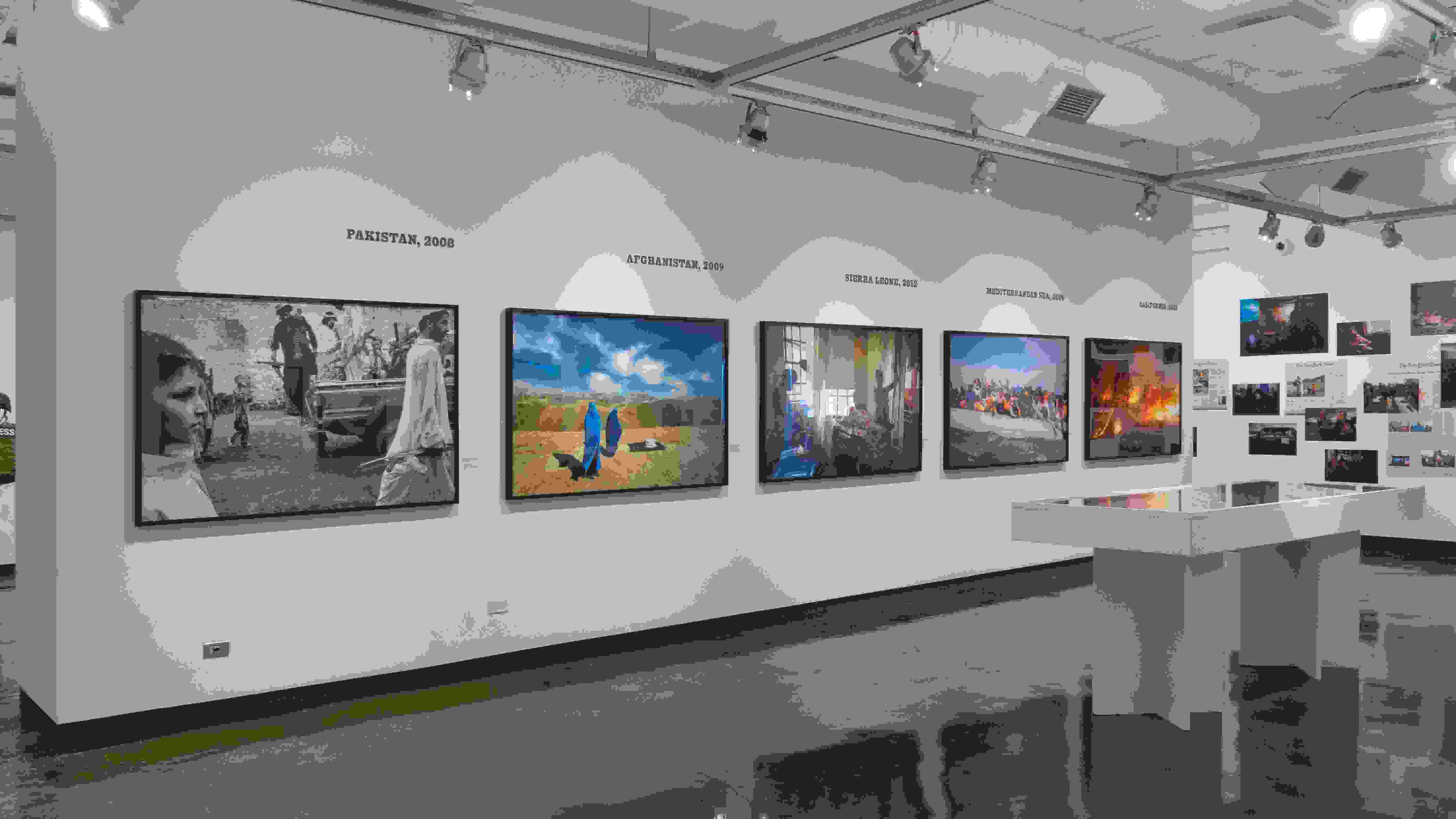Photographs depicting war and conflict are hung across two gallery walls. In the center of the space is a large vitrine with ephemera inside.