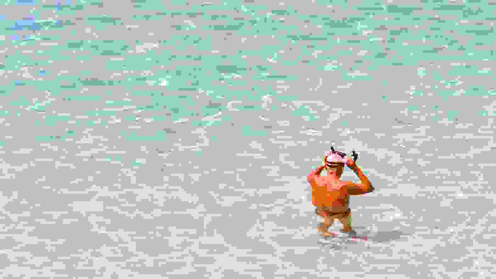 Sun drenched photo of an open sea landscape; in the bottom right corner, an older person is putting on goggles, seemingly about to go for a dip