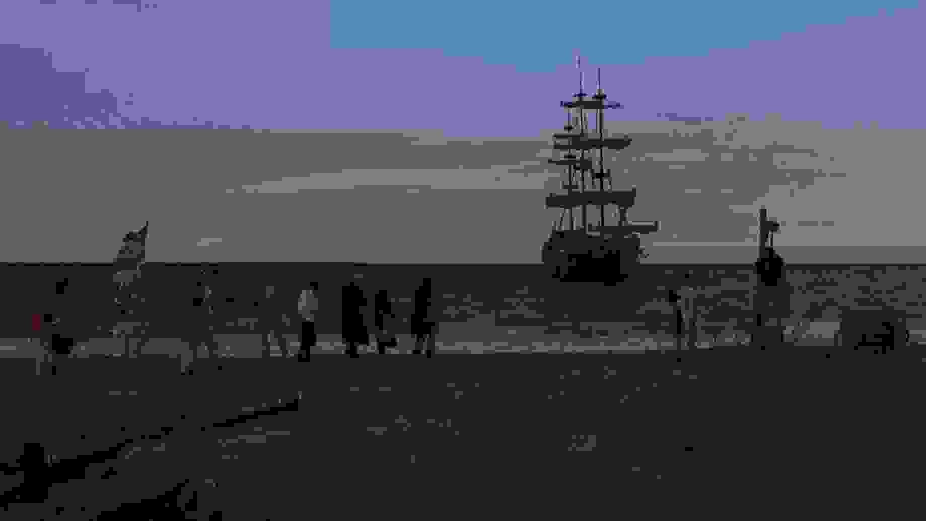 Dramatic sunset frame of a ship near shore; on the beach, a group of people walk, one carrying a union jack flag