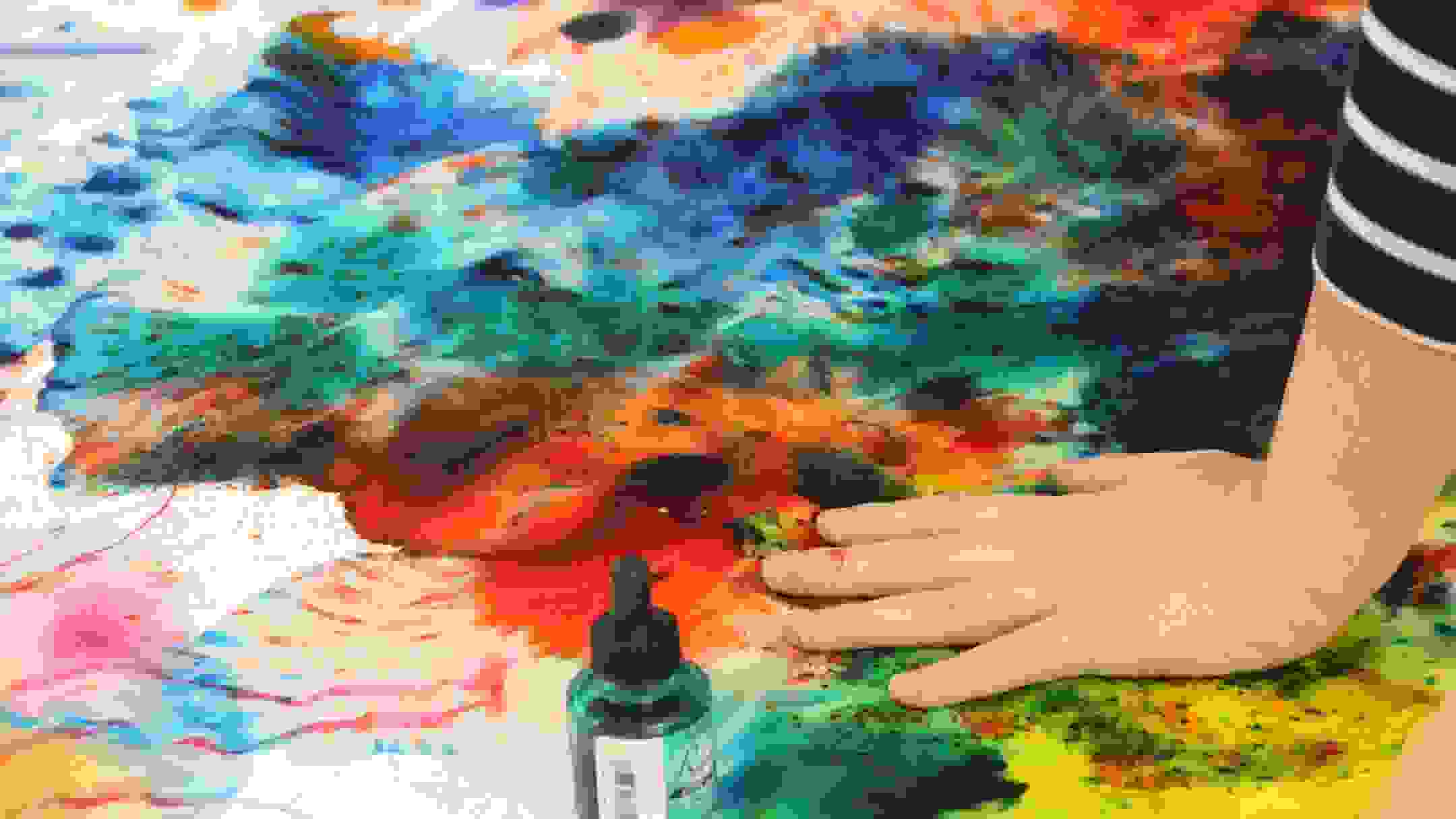 Photograph of a multicolored canvas on the floor with a hand atop supporting the weight of someone in the act of painting it
