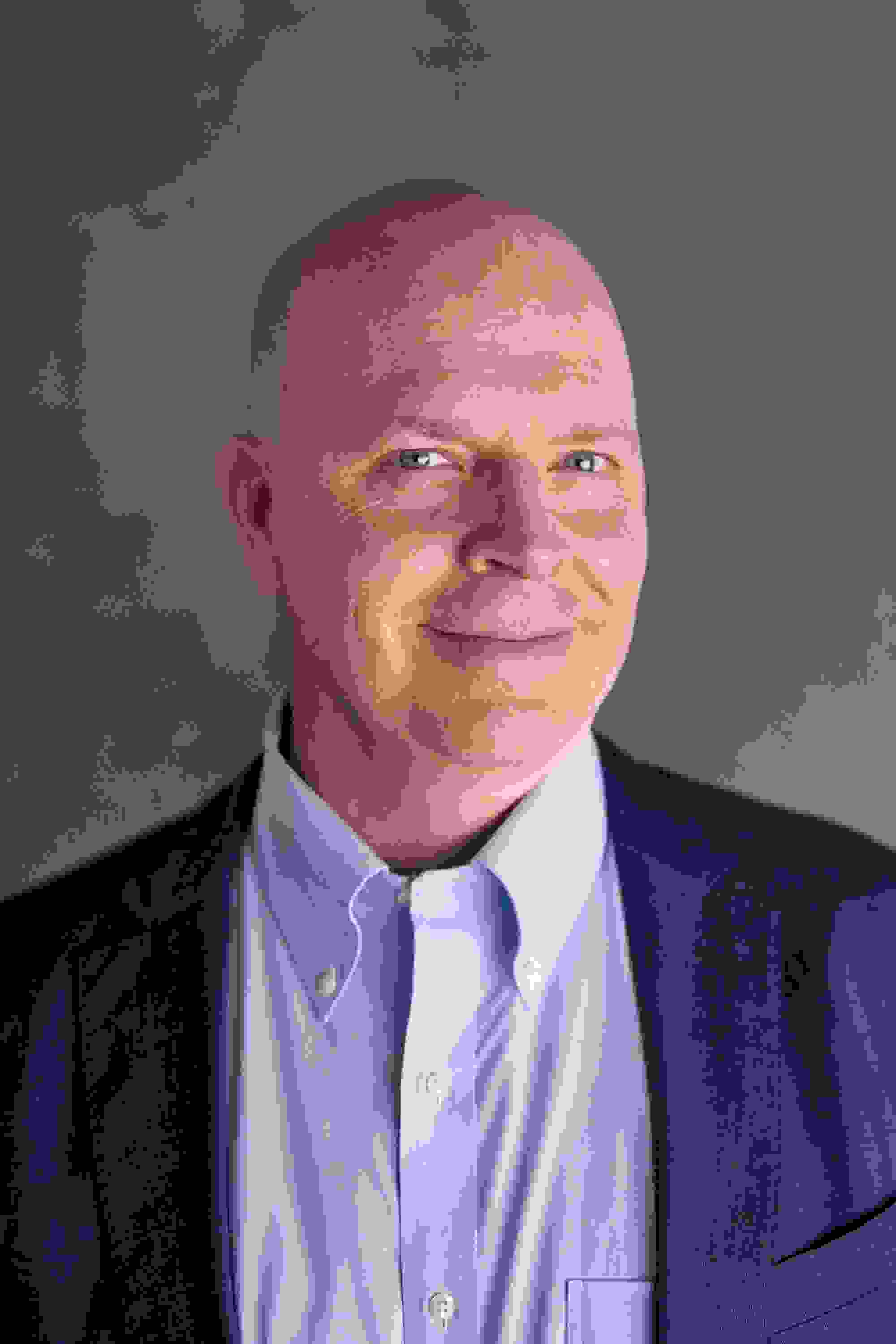 A middle-aged man with light skin and blue eyes, wearing a light blue button-down shirt and a navy blazer. He is smiling slightly and looking directly at the camera. The background is a neutral gray, creating a professional and approachable tone.