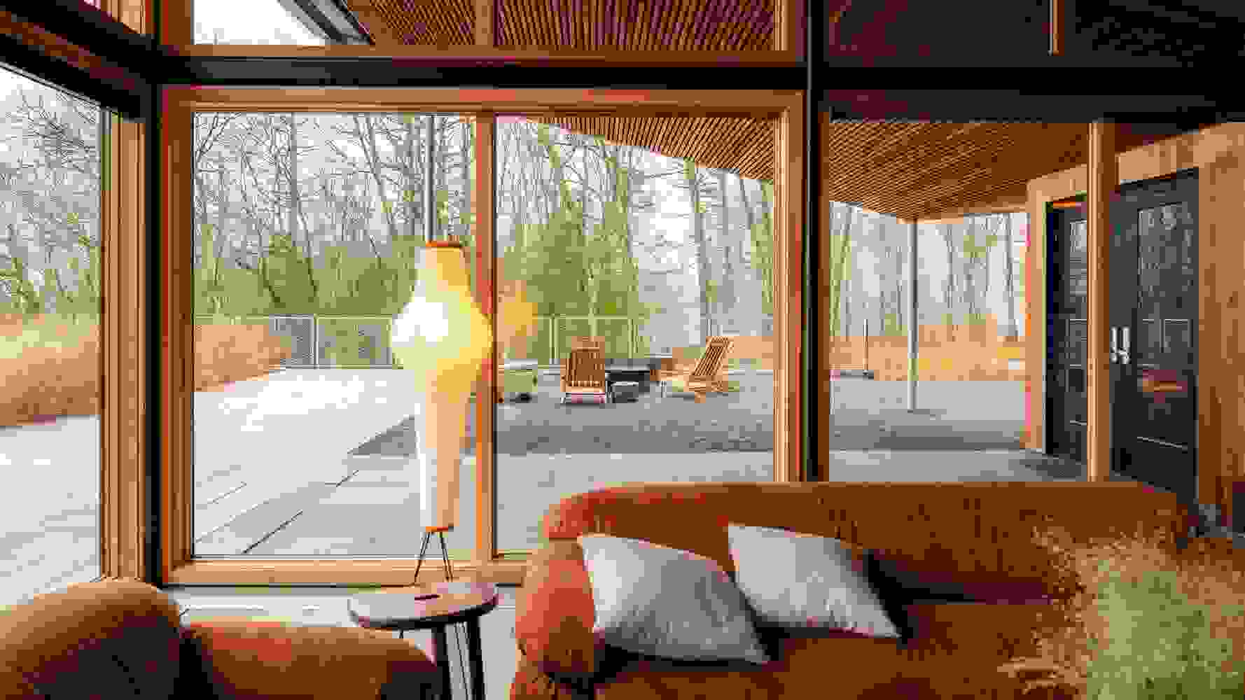 The interior of a wooden style resort reception area with a forest view. The overall color is warm orange. The foreground features two dark orange minimalist retro style matte fabric sofas, with a chocolate colored mini side table in between.