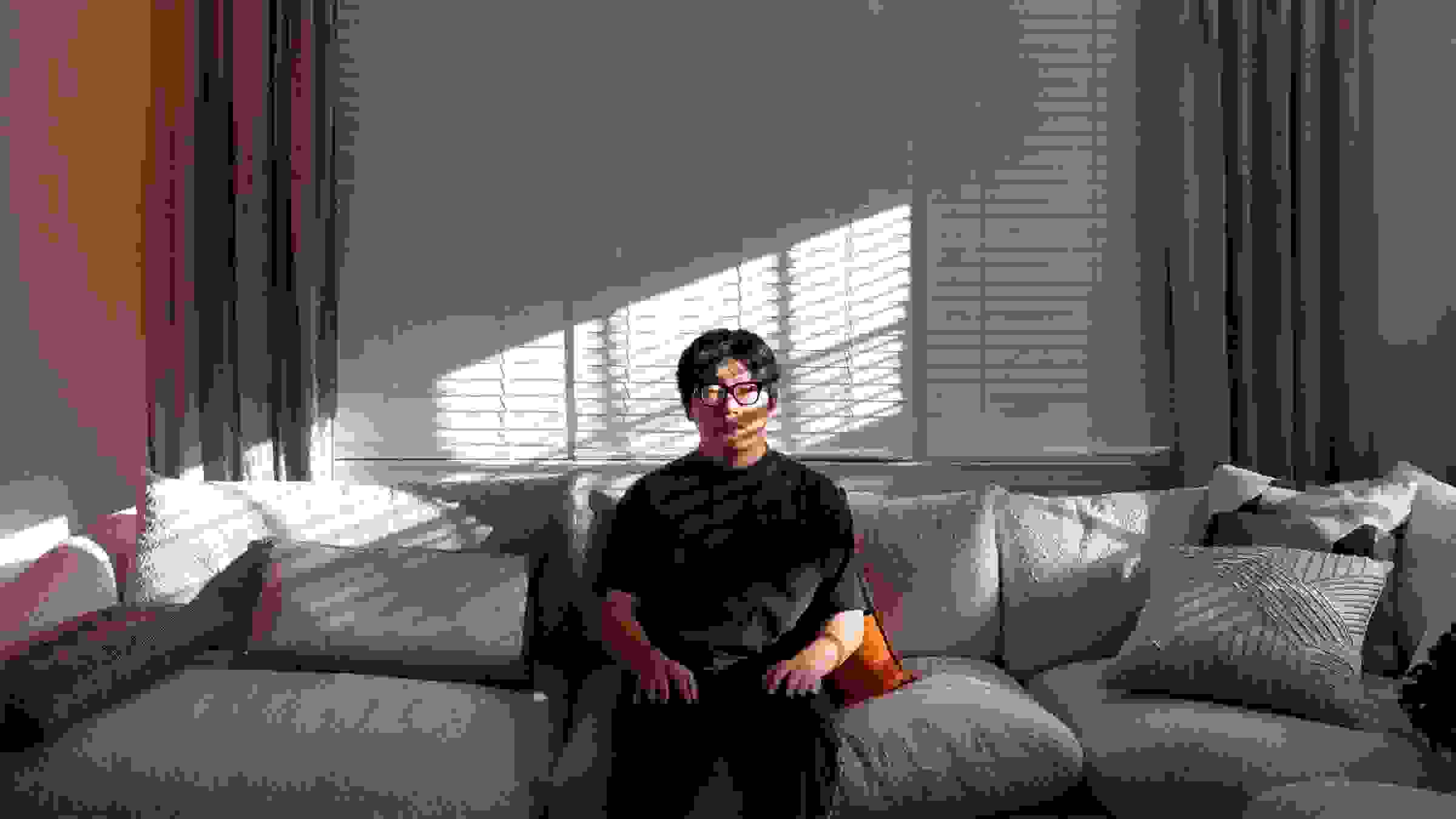 In this color photograph, the artist sits at a couch while looking into the camera. Behind him are drawn curtains.