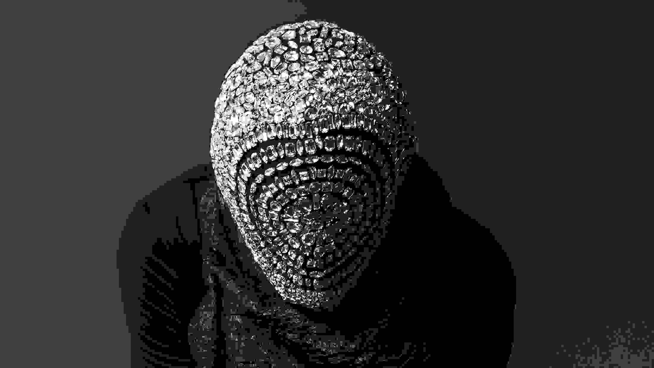 A black-and-white studio fashion photograph of a person wearing a diamond mask and black dress.