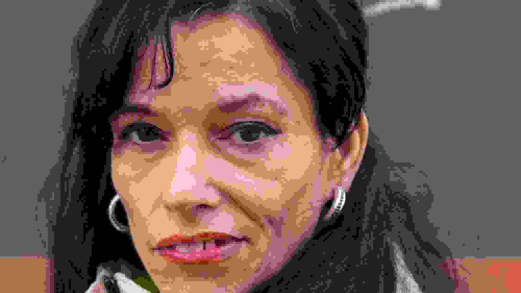 A documentary portrait photograph of a middle-aged woman with dark hair. She is wearing a white jacket and red lipstick and looking at the camera with a smile.