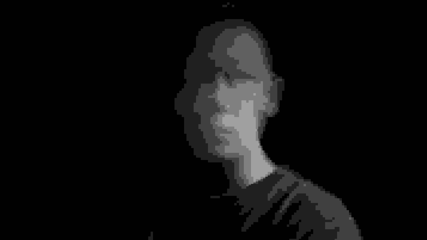 A black-and-white self-portrait of a man sitting in front of a black background. His face is slightly blurred and he is looking at the camera, and he is mostly in shadow.