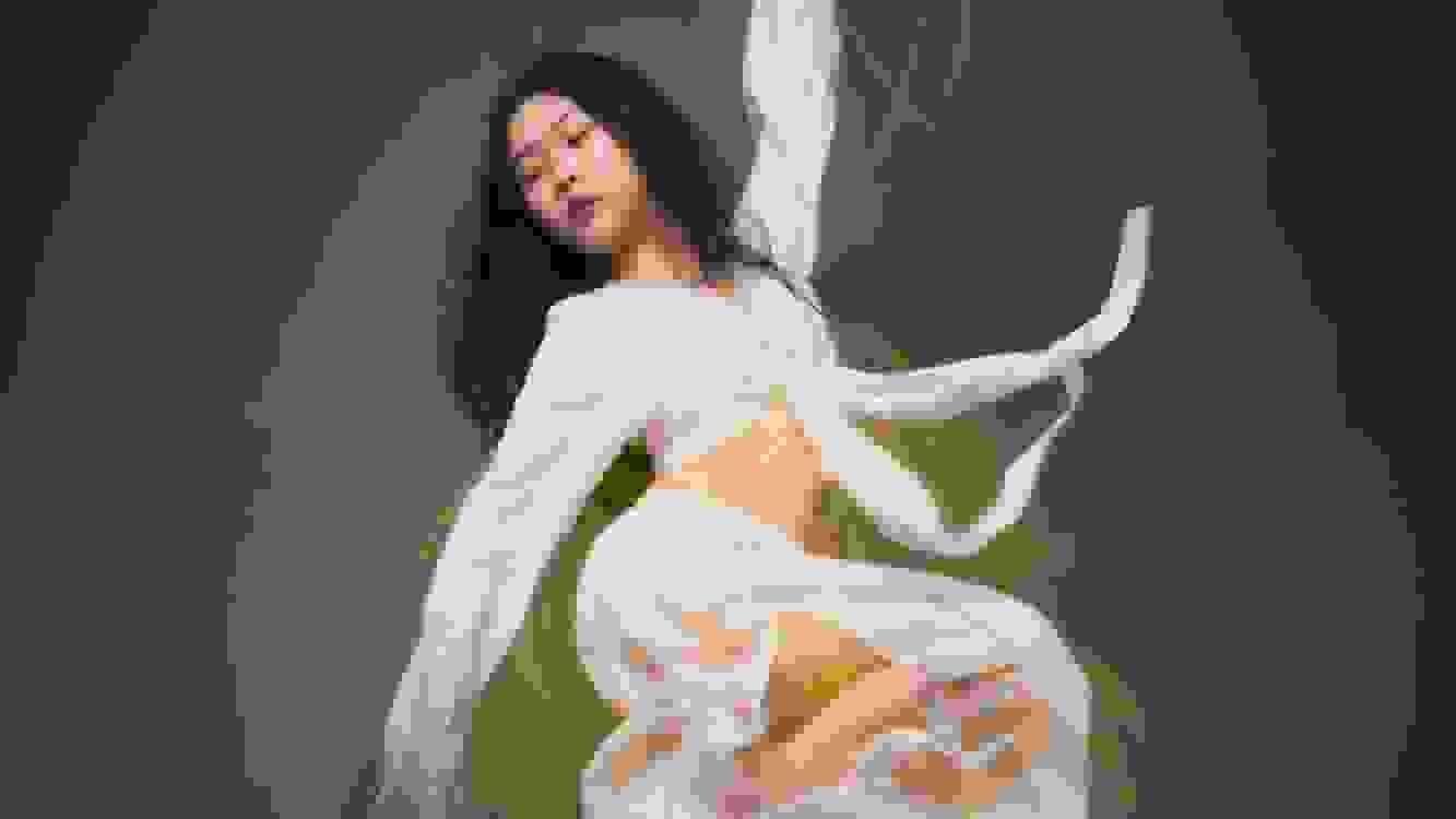 A photograph of a woman in mid-dance pose. She is wearing all white and is holding two pieces of fabric which are in motion. There are yellow streaks of yellow light indicating her movement around her.
