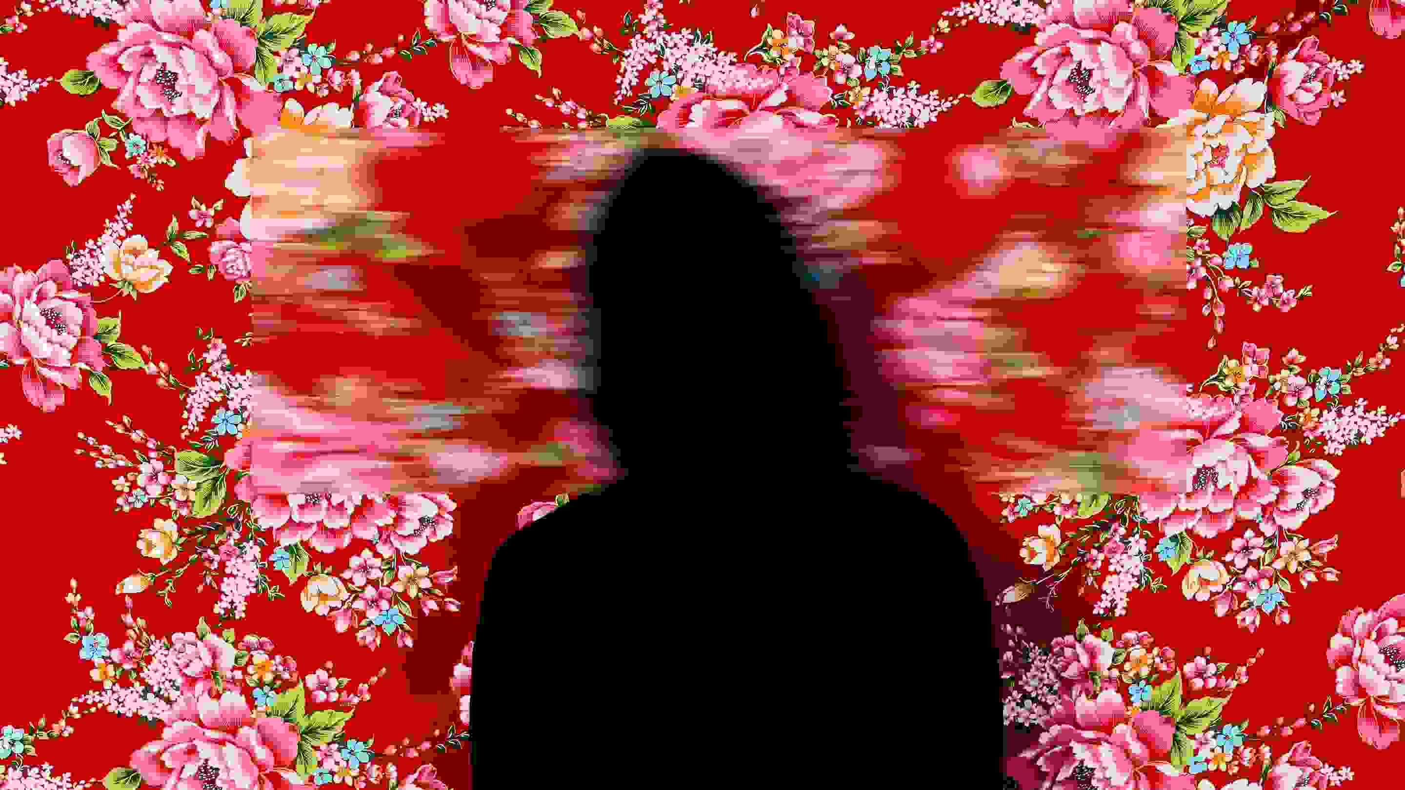 A color photograph featuring a figure wrapped in black from head to toe standing facing the camera against a red Hakka floral fabric background, which is suspended and reveals a faint dark wooden floor below. Three blurred rectangles cover the head, hands, and feet.