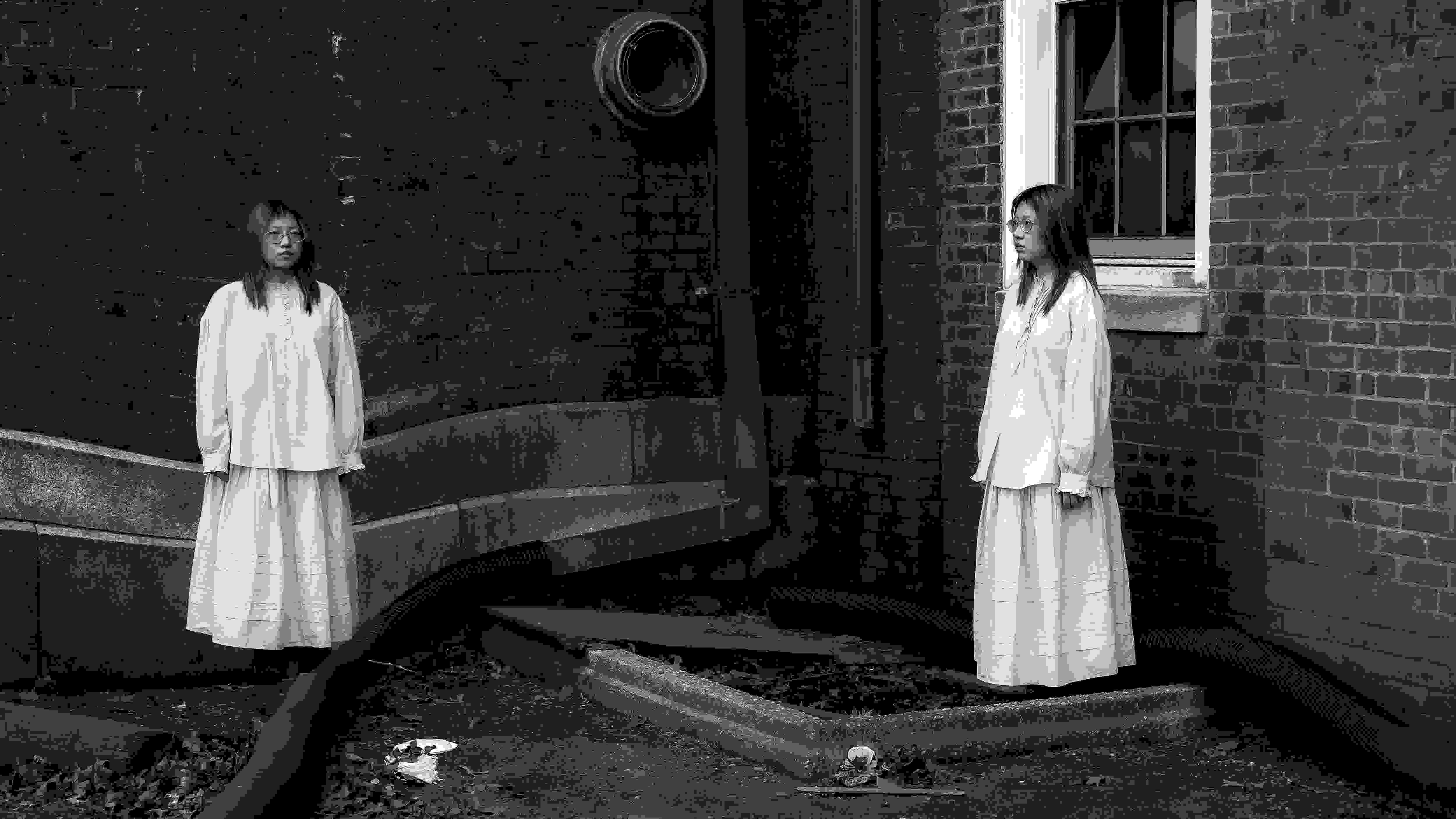 Black and white photograph of a girl duplicated in the frame, standing at two corners of a brick building. Both wear the same long dress, facing each other across a barren, debris-littered ground with industrial pipes.