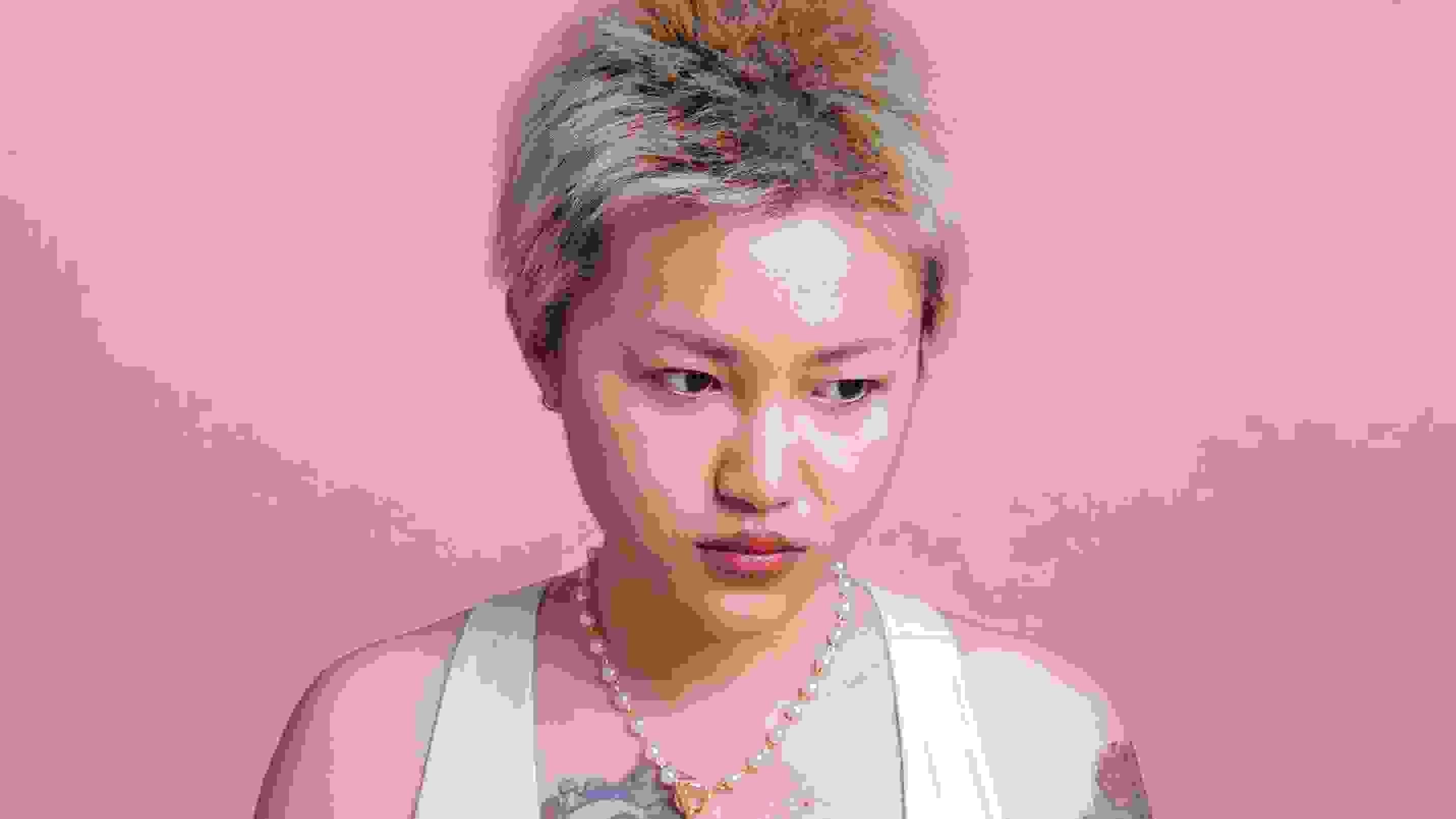 In this color photograph: On a pink background, a half-length portrait of a gold-haired young Asian woman wearing a white sports bra, sitting. She has large colorful tattoos on her right chest and left arm. On the pink table in front of her are three red  tomatoes. She is holding a smaller dark- colored tomato and is looking away  from the camera, appearing thoughtful.