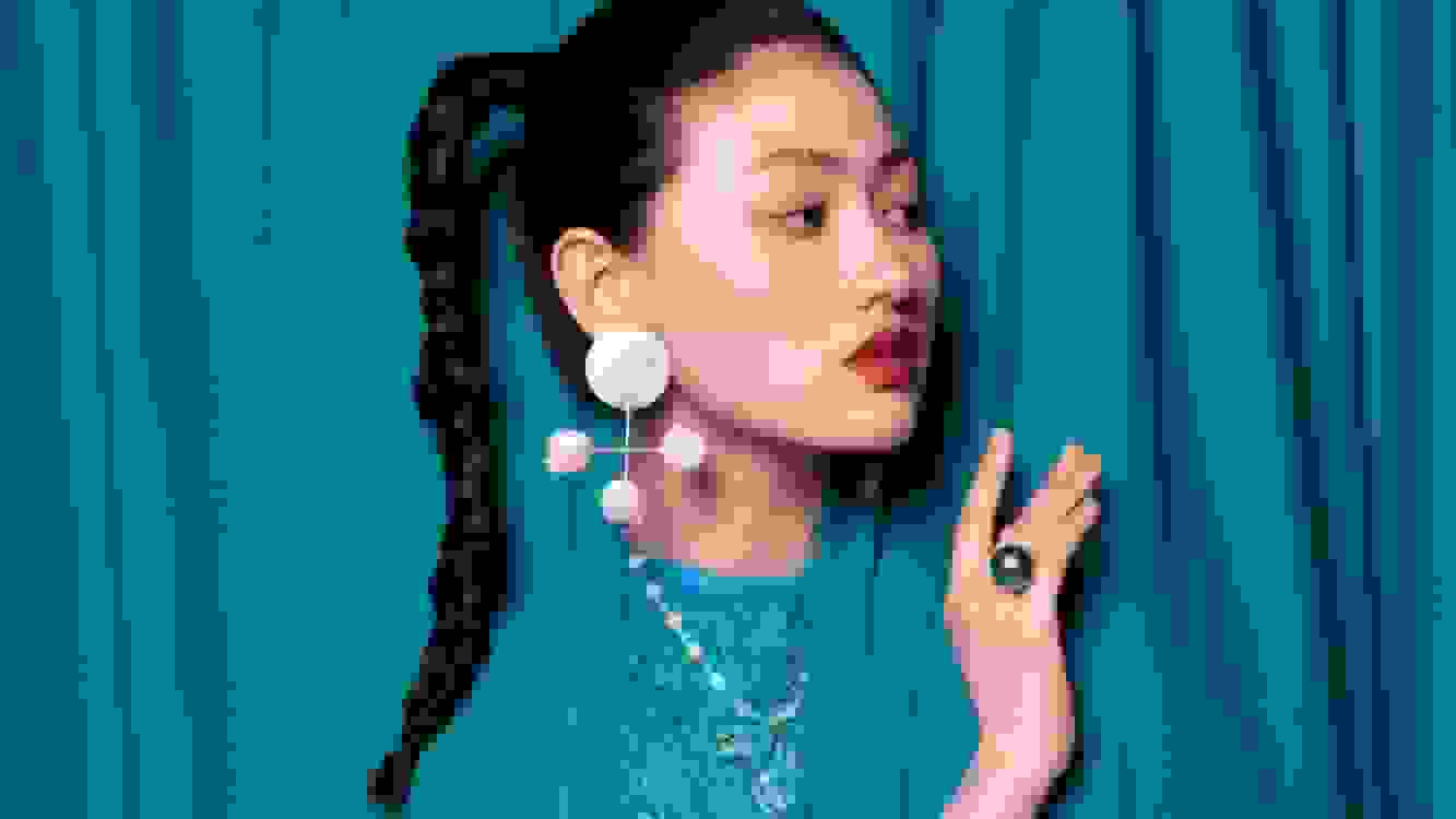 A photograph of a woman wearing a blue ruffled top peeking her head and arm out from behind a curtain of the same color. She is looking off-camera to the right and has her hair in a braid, and is wearing large white earrings, red lipstick, and a large ring on her middle finger.