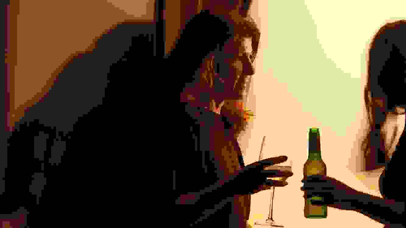 A color photograph of two women by a large open window at nighttime. They are in conversation and one woman holds a wine glass while the other holds a beer bottle.