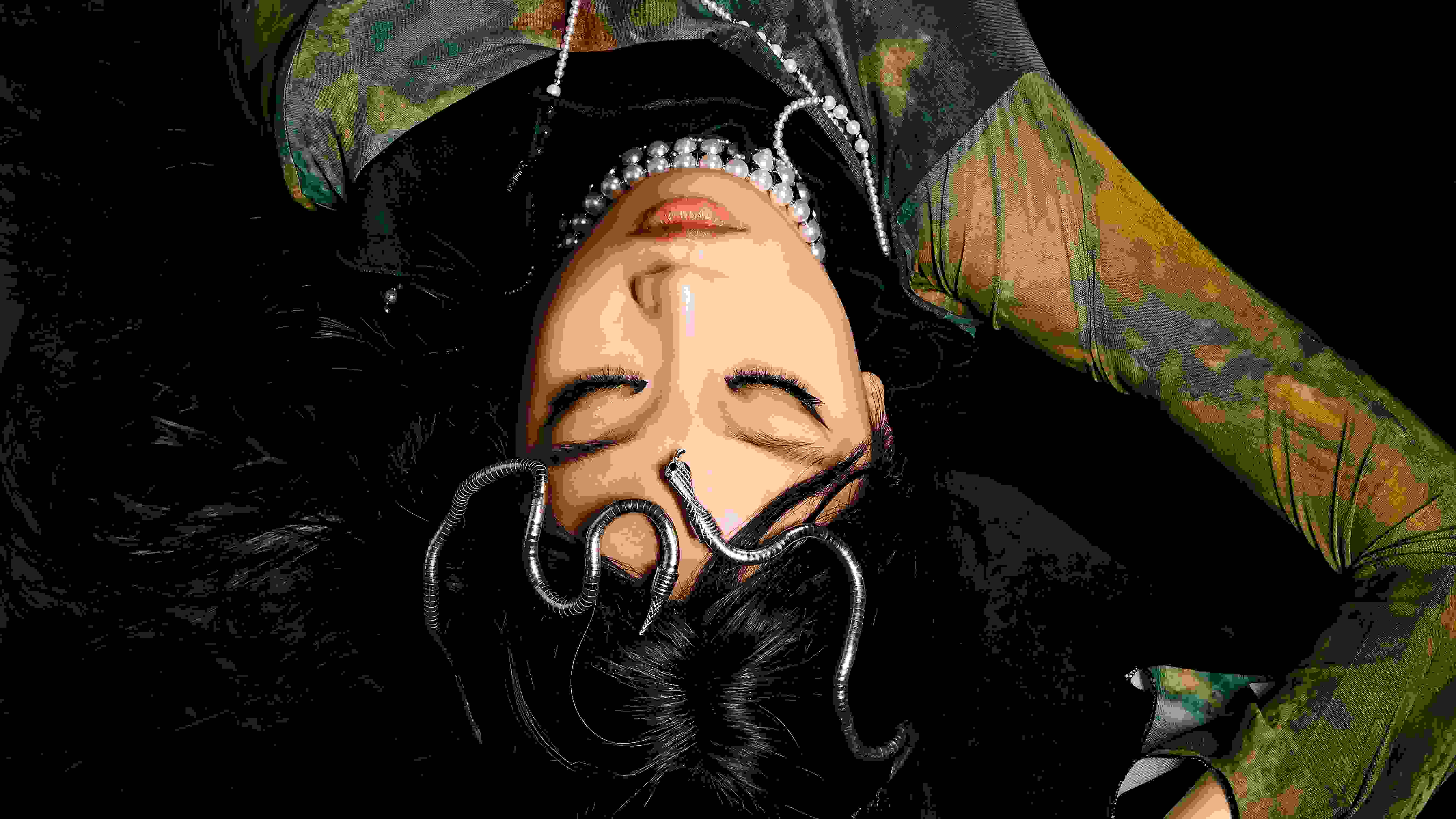 A woman lays on a black background, wearing a green long sleeve dress and a snake headpiece. Her eyes are closed and there is a skeleton of a snake slithering up her chest.