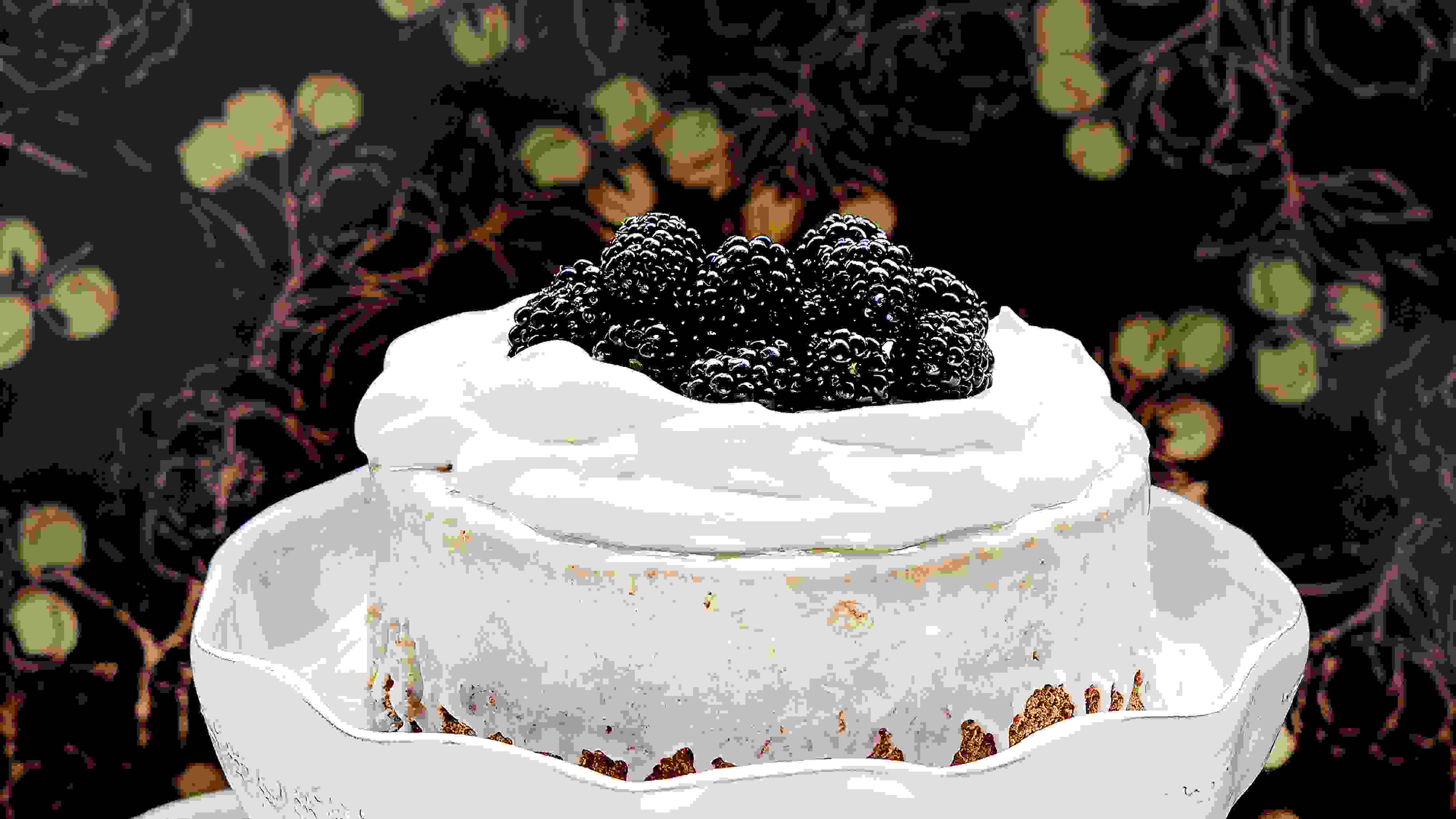 A cake that is covered with sugar glaze and topped with whipped cream and blackberries on a white cake stand. The background it dark with a gold pattern and there are Christmas ornaments and a silver fork next to the cake stand.