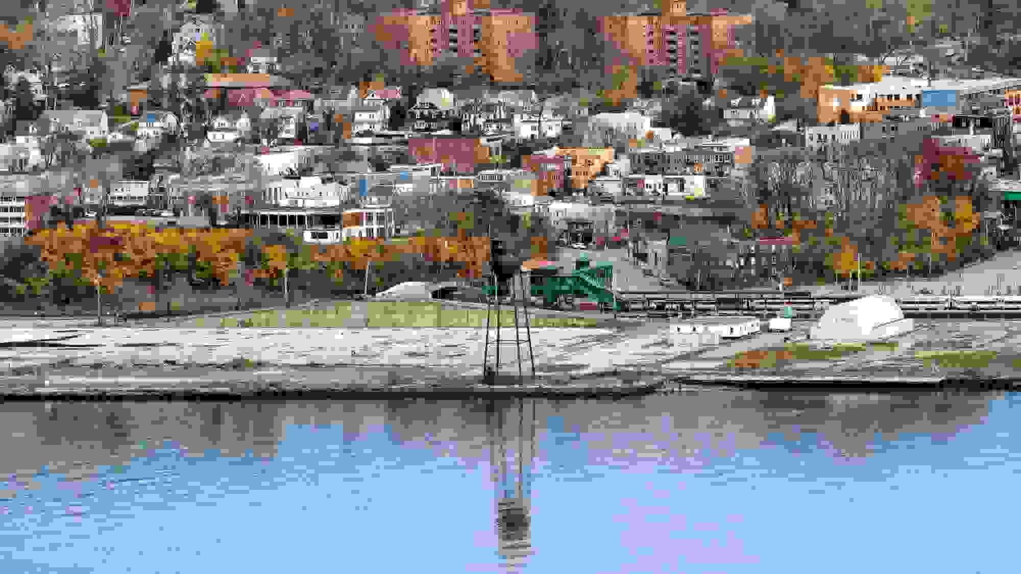 A documentary photograph of a water tower, trees, and buildings, near the shoreline of a river.