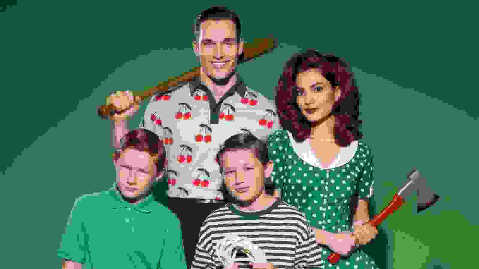 A formal studio portrait of a family of four, including a dad, mom, and two kids. Each of them is holding a different weapon. They are all wearing preppy, patterned clothing and standing in front of a green background.