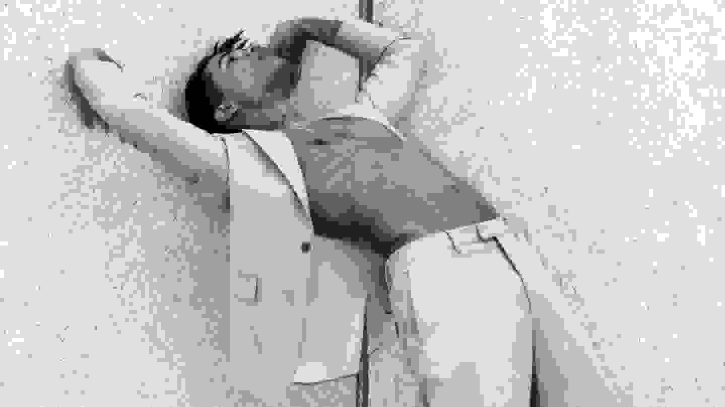 A black-and-white photograph of a man in a suit and dark shoes doing a backbend against a wall. The front of his jacket is open and exposing his chest.