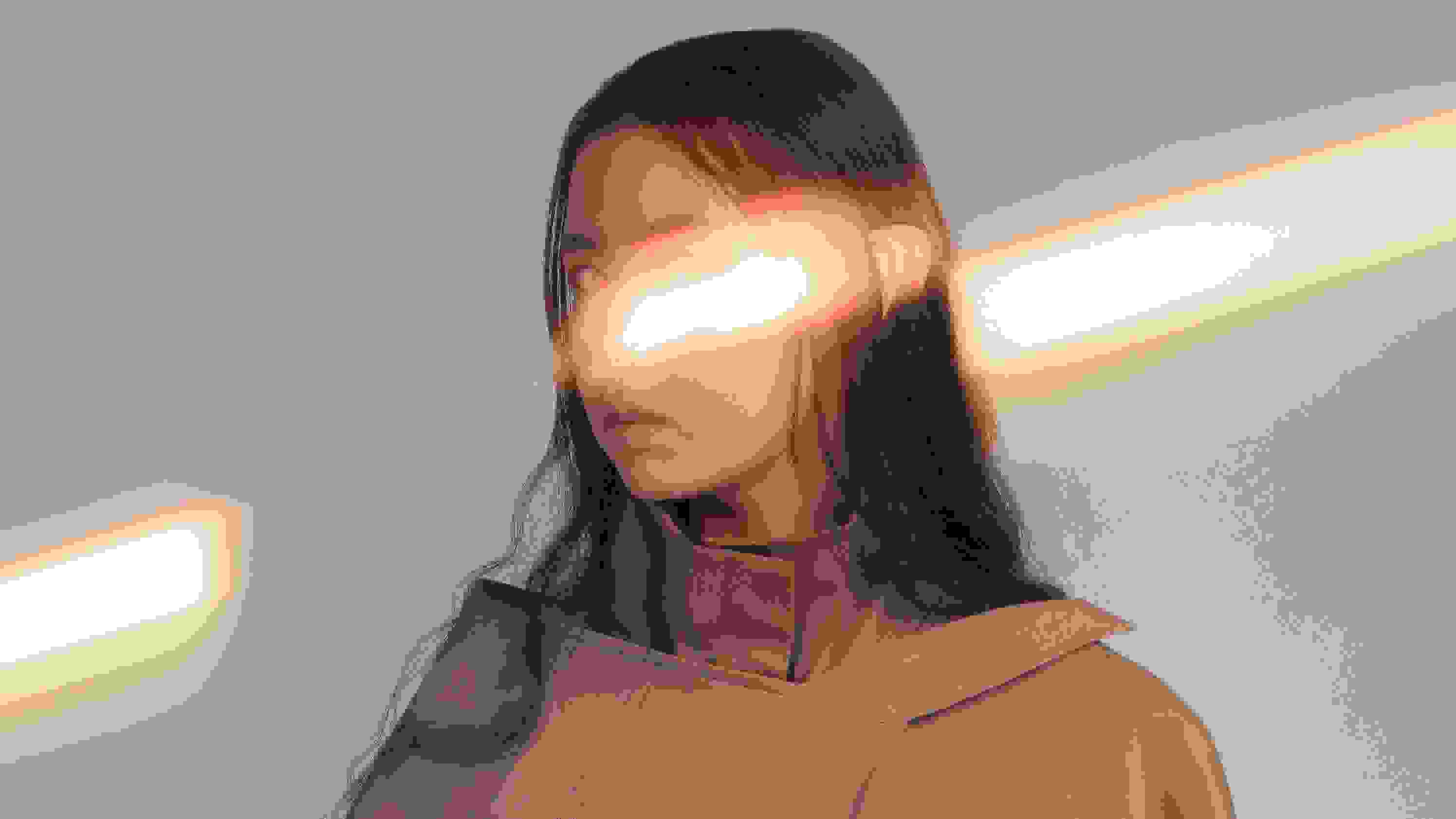 A photograph of a woman with dark brown hair in front of a tan background. She is wearing a tan jacket covered in buckles and pockets. SHe is looking at the camera. There is a streak of light going across the frame and over the model’s face, covering her nose and left eye.