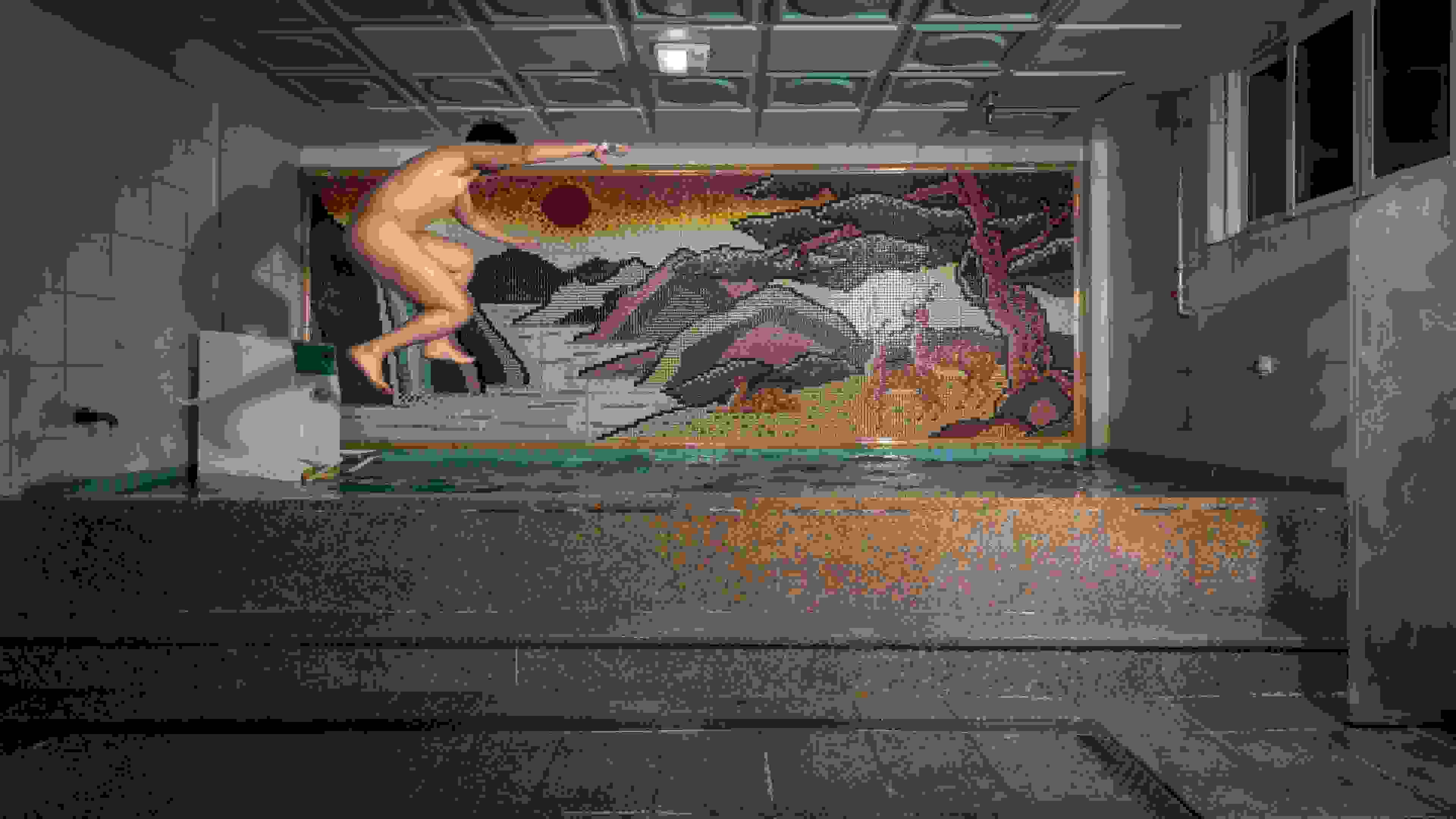 A documentary photograph of a nude man in profile, jumping from left to right side of a large tiled bath into the water. On the wall behind him is a colorful tile mosaic of a landscape.