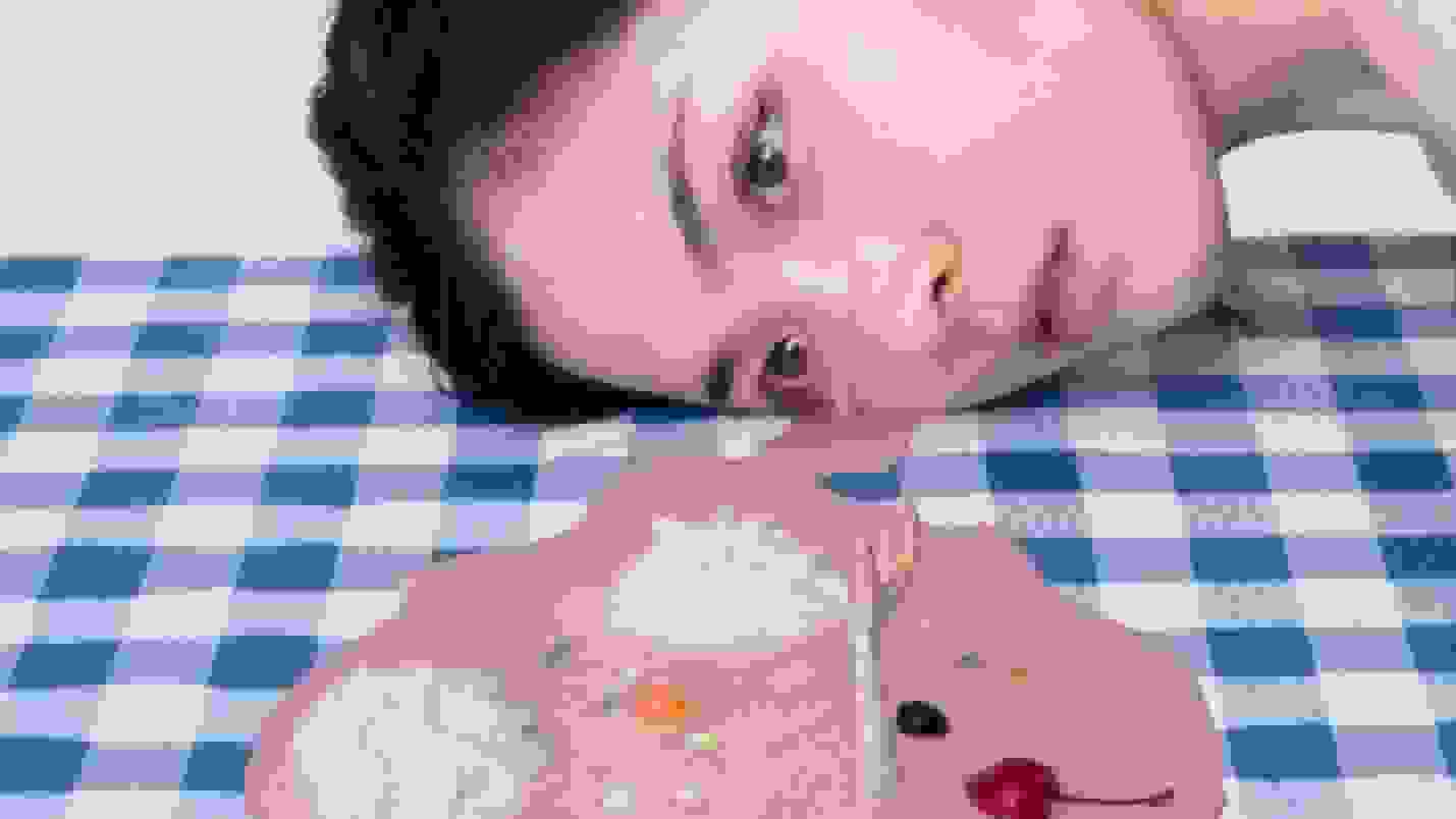 A digitally-manipulated photograph of a table covered with a blue and white checkered cloth. A girl is resting her head on it and crying a tear of melted ice cream. On the table in front of her is a puddle of ice cream with cherries and whipped cream. There is an arm with a hand reaching upwards sticking out of it.