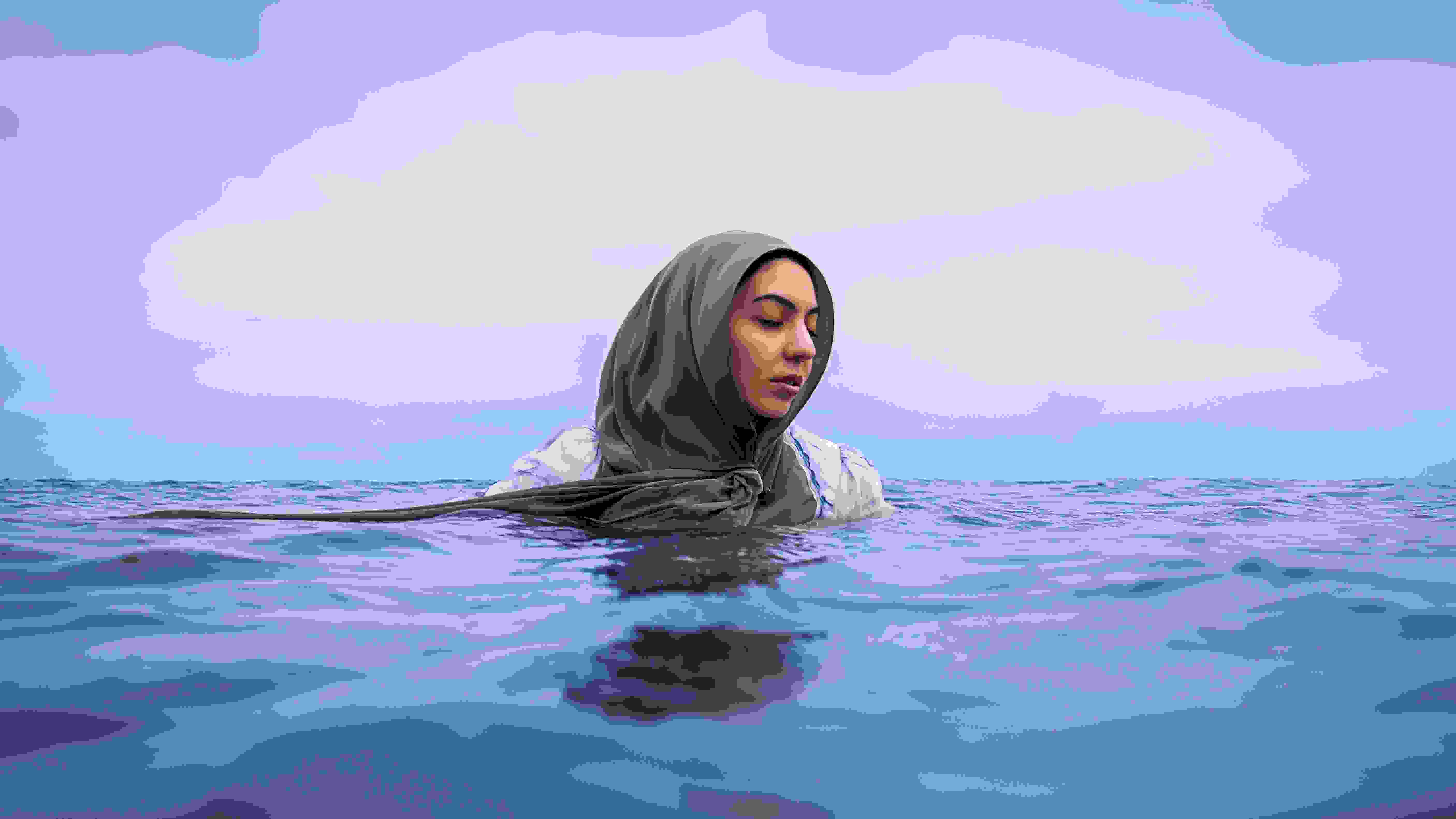 A woman wears a light purple hijab and is partially submerged in calm ocean waters. The horizon line divides the frame, with the vast, tranquil sky above and the gently rippling water below. The woman has a serene and contemplative expression, her eyes closed as she floats.