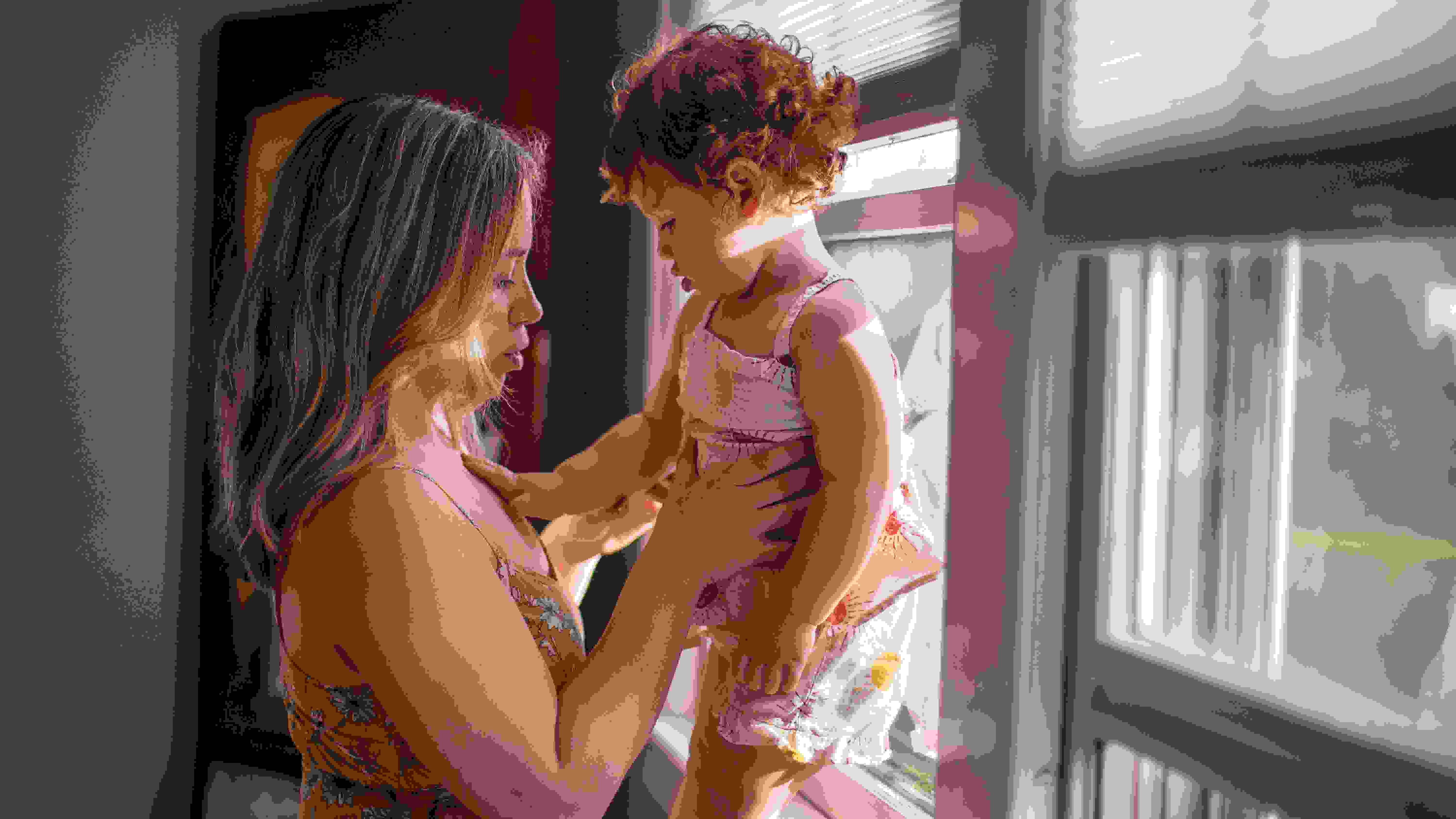 A woman holding her baby who is standing up next to the window, the baby’s hand in touching the woman’s chest. They both are looking at the baby’s hand. The woman is wearing a floral dress and the baby a floral jumpsuit. Her legs are not visible.