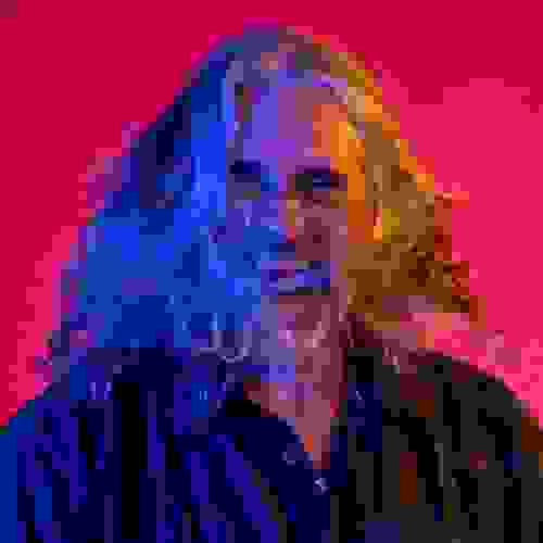 A portrait of Michael Kaminski in front of a hot pink backdrop. Michael has long, silver hair, which in the picture appears to be spun around. His face, along with his hair, is lit by a blue light on the left and an orange light on the right. 