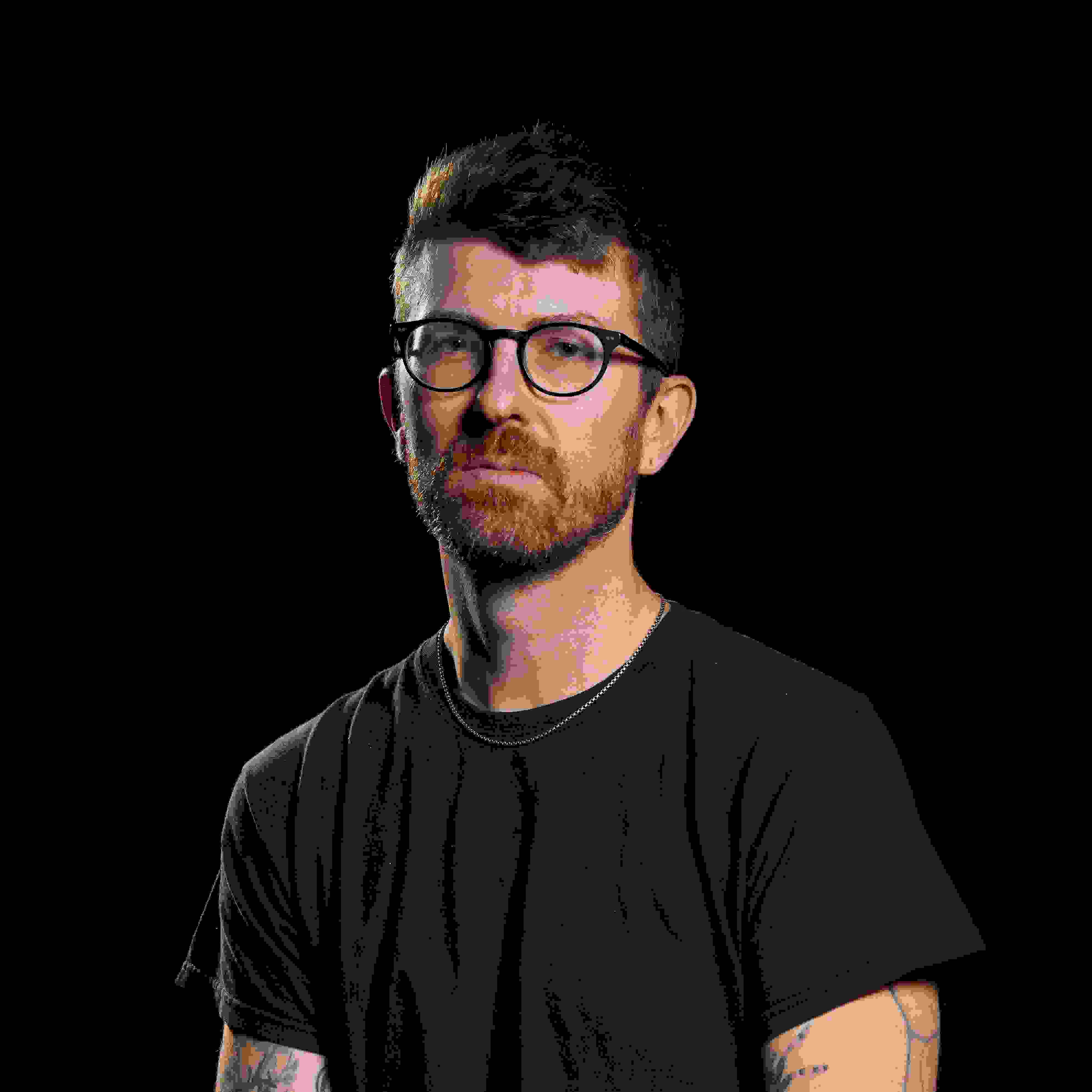 portrait of a man in a studio environment with a dark background wearing a black tshirt. He is wearing glasses and has a silver necklace. 