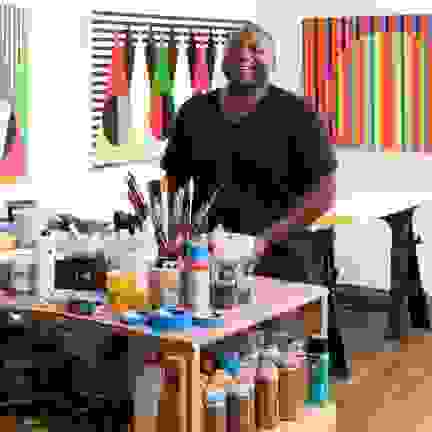 Rico Gatson in a black t-shirt smiles in his studio, surrounded by bold, geometric paintings. 