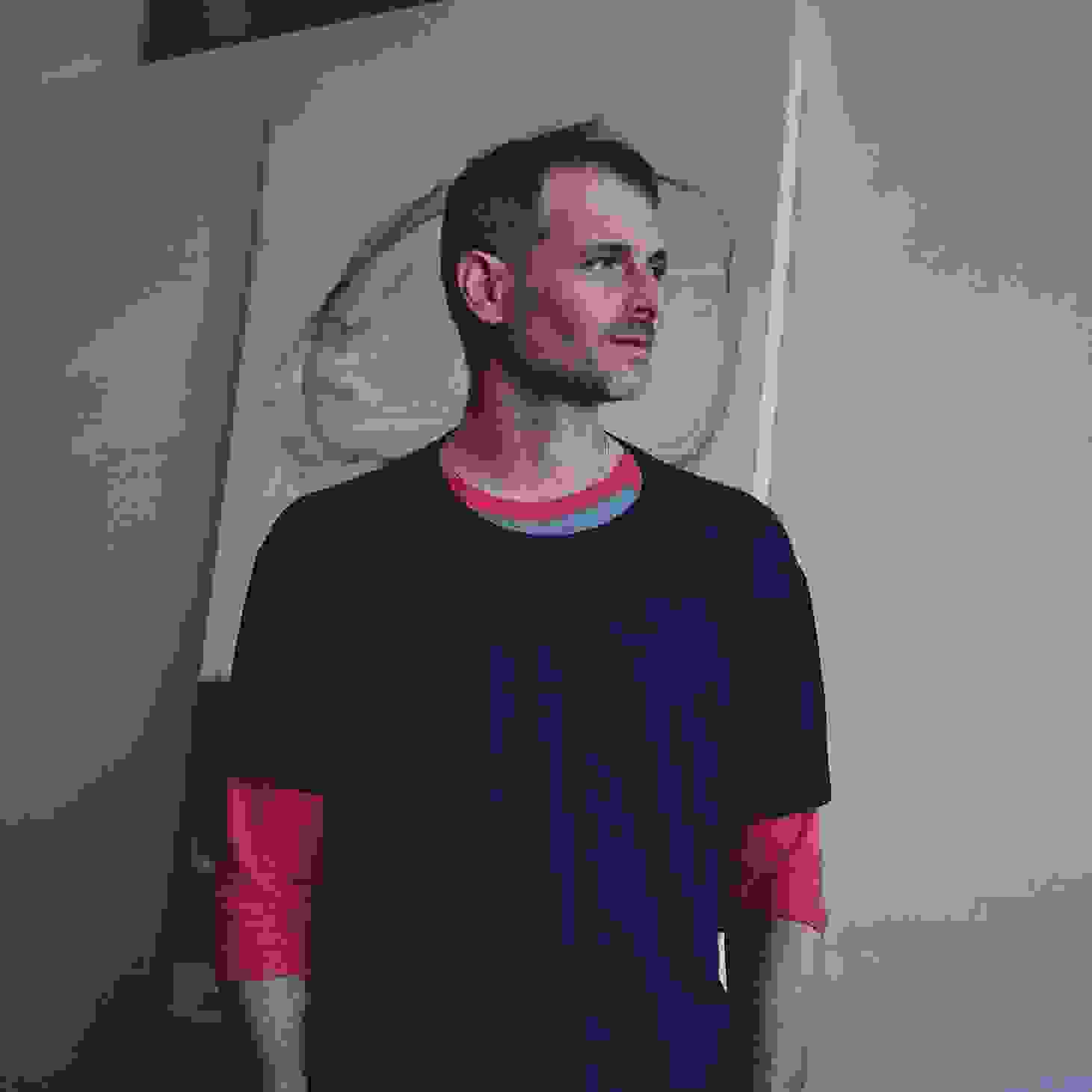 Uri Aran in a navy t-shirt and red long sleeves stands in an art studio, gazing to the side, with a large abstract canvas behind him.