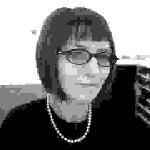 A black and white photo of Debra Klomp Ching, who is looking into the camera with her head slightly turned to the right. She has light skin, short hair, and is wearing a pair of glasses with black frame. She is wearing a black buttoned blouse and a pearl necklace. 