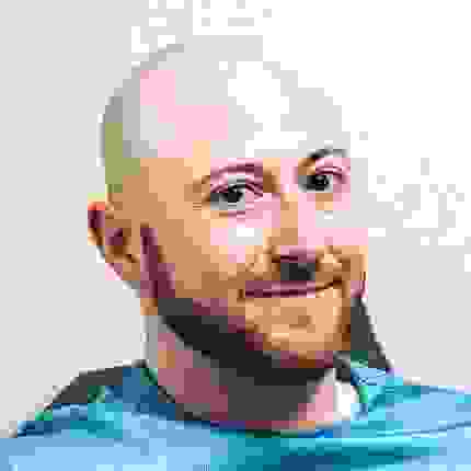 Kris Grey  smiles while wearing a blue shirt, sitting against a neutral background.